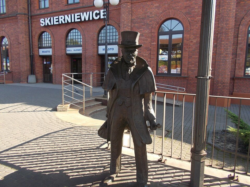 Zdjęcie przedstawia peron dworca kolejowego w Skierniewicach współcześnie. Na pierwszym planie widoczny jest pomnik Stanisława Wokulskiego. Ukazana jest męska postać w garniturze, płaszczu i cylindrze. Na drugim planie widać ceglany budynek dworca i napis Skierniewice.