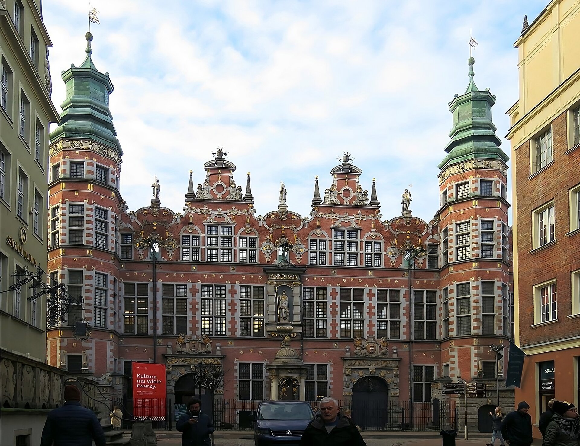 Zdjęcie przedstawia widok na Wielką Zbrojownię od strony ul. Piwnej w Gdańsku. Obiekt utrzymany w stylu manieryzmu niderlandzkiego, widoczny parter budynku oraz dwie kondygnacje, w tym ostatnia ze szczytem budynku. Kondygnacje na piętrach mają wpasowane w konstrukcję fasady okna prostokątne lub, w przypadku najwyższej kondygnacji, okna wpasowane w obramowanie łuków pełnych. Zdwojenie elementów poprzez dwa portale wejściowe z herbami miasta na parterze, dwa szczyty osłaniające dach budynku i dwie flankujące budynek wieże wieloboczne z oknami. Obiekt kunsztownie zdobiony przy pomocy złoceń na portalach i pod fryzami budynków, na szczytach budynku widoczne sterczyny, woluty i ornamenty okuciowe. Wieże wykończone hełmami. Pośrodku konstrukcji, na piętrze w niszy, figura alegoryczna przedstawiająca boginię Artemidę. U dołu fotografii kilkoro przechodniów i samochód osobowy.