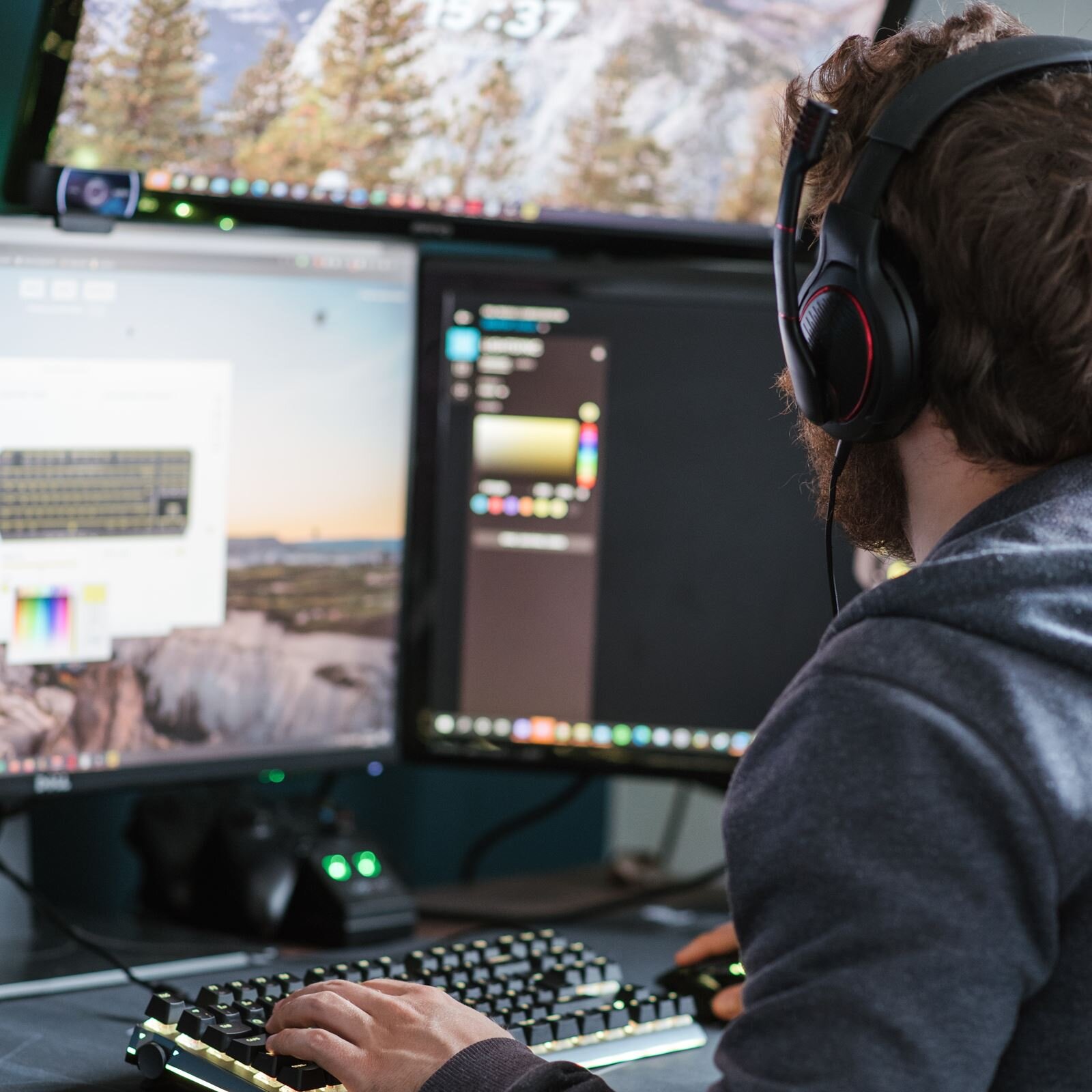 Zdjęcie przedstawia postać mężczyzny, od pasa w górę, ukazaną od tyłu, siedzącą przy biurku, przodem do monitorów komputerów. Na biurku widoczne są dwa monitory komputerów stacjonarnych oraz klawiatura, na której spoczywa lewa dłoń mężczyzny, i myszka, na której spoczywa prawa dłoń mężczyzny. Mężczyzna na uszach ma słuchawki. Na monitorze po lewej wyświetla się klawiatura, zaś na monitorze po prawej widoczny jest rozmyty panel z ikonami. 