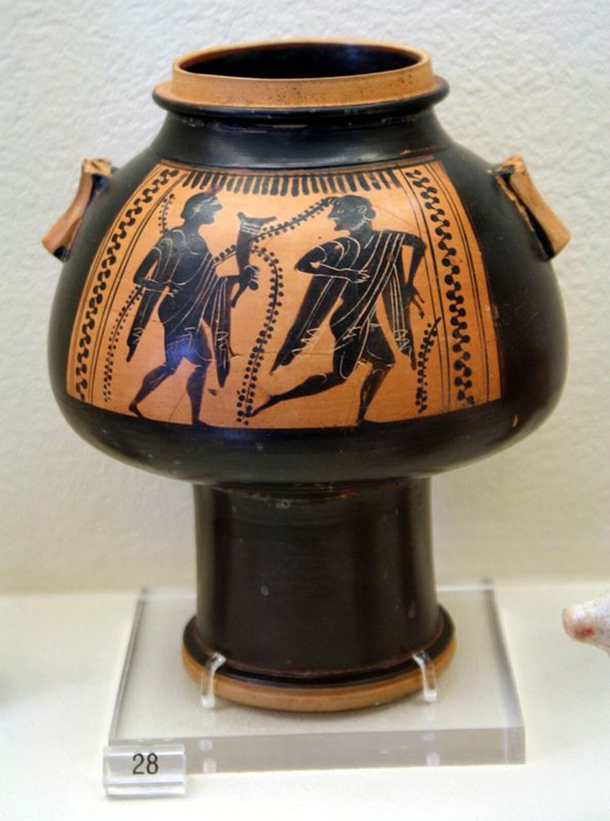 Ilustracja przedstawia naczynie greckie o nazwie psykter. Przedstawia dwie postacie - pierwsza biegnie, oglądając się, druga idzie, trzymając róg. Po bokach są uszy.