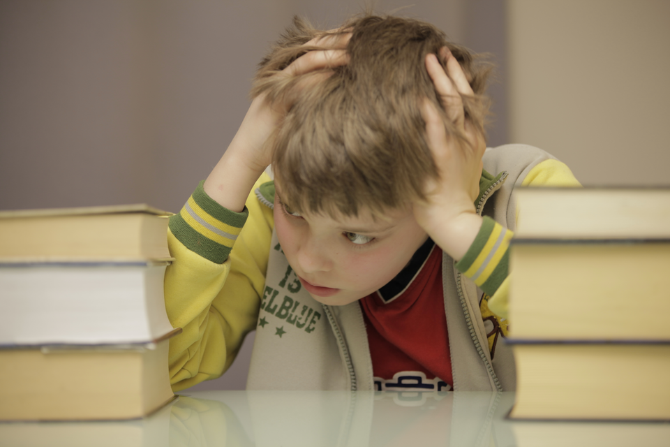 Fotografia przedstawia zestresowanego, zmartwionego chłopca. Chłopiec siedzi przy biurku, opiera na nim łokcie i podpiera głowę dwiema rękami. Na biurku, po obu jego stronach, znajdują się stosy książek.