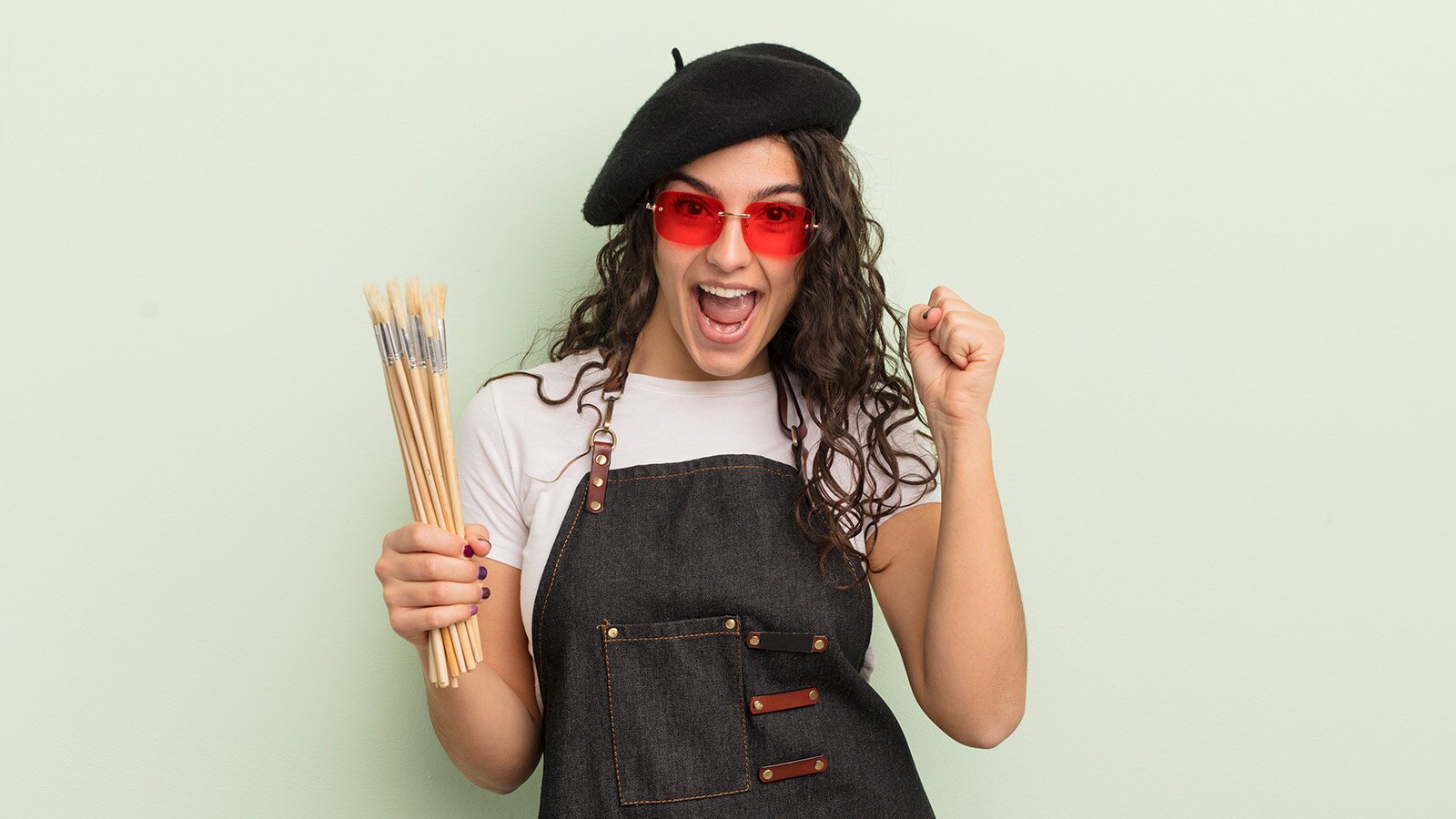 Zdjęcie przedstawia młodą kobietę, w ujęciu od pasa w górę. Kobieta ma długie, ciemne i lekko kręcone włosy, na które założyła czarny beret. Jest ubrana w białą bluzkę z krótkim rękawem i dżinsowy fartuch. Na twarzy ma czerwone okulary. Obie ręce ma podniesione do góry i zgięte w łokciach. W prawej dłoni trzyma pęk cienkich pędzli, a lewą dłoń ma zaciśniętą. Szeroko się uśmiecha.