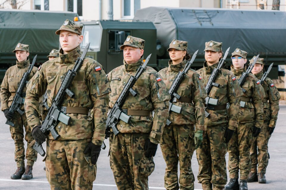 Zdjęcie przedstawią grupę żołnierzy stojących na baczności. Są oni ubrani w mundury armii polskiej, Z przodu mają przewieszone karabiny. Z tyłu za nimi widać pojazdy wojskowe.