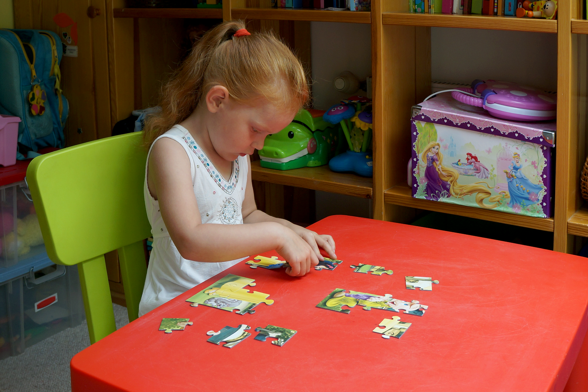 Fotografia przedstawia dziecko w wieku około czterech lat. Dziewczynka siedzi przy czerwonym stole, samodzielnie układa puzzle. W tle znajduje się drewniany regał z licznymi zabawkami.