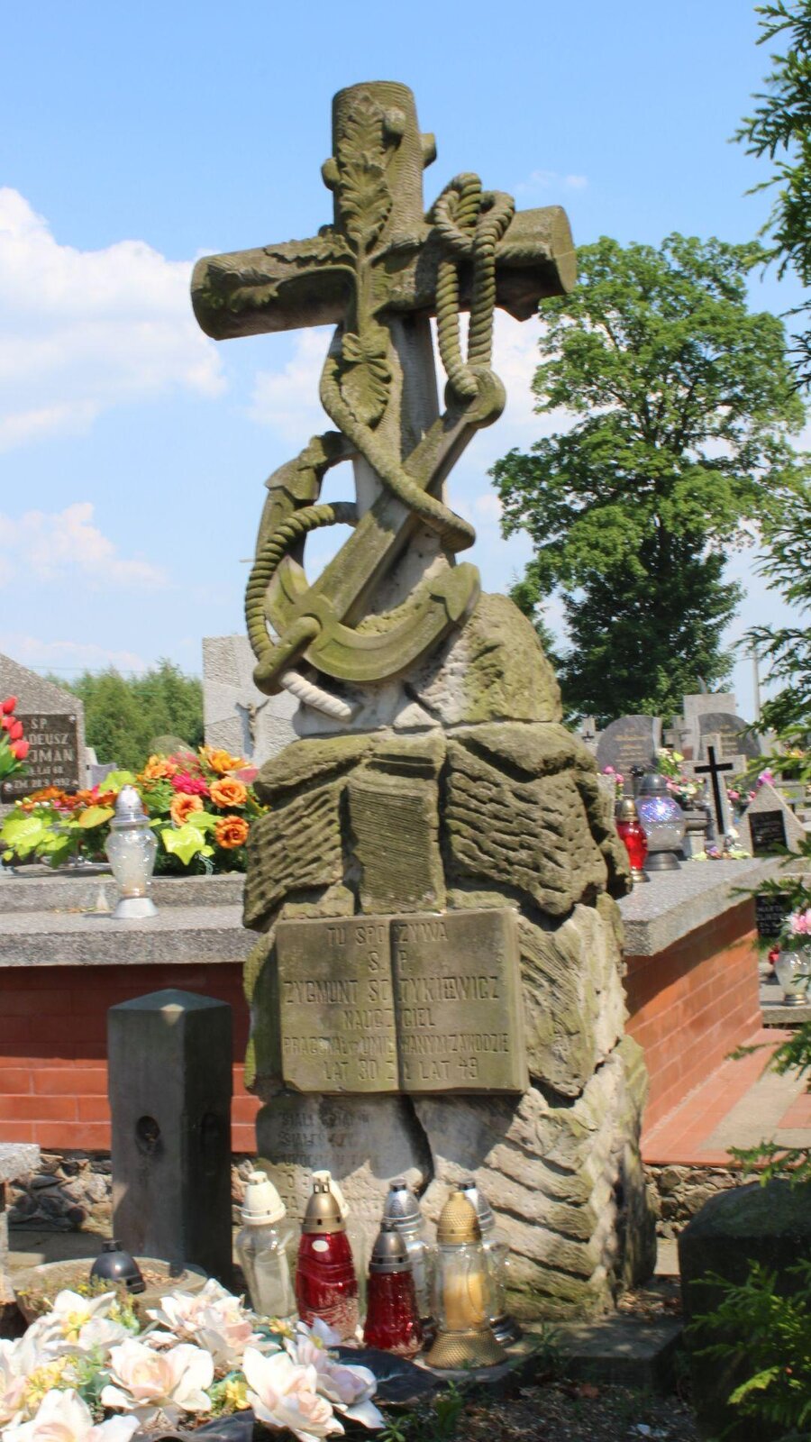 Zdjęcie przedstawia wysoki, kamienny nagrobek, na czubku którego został umieszczony krzyż obwieszony kotwicą na sznurze. Poniżej znajduje się tablica pamiątkowa z napisem "TU SPOCZYWA ŚWIĘTEJ PAMIĘCI ZYGMUNT SOŁTYKIEWICZ, NAUCZYCIEL". Reszta napisów nieczytelna. Pod pomnikiem stoją białe, srebrne i czerwone znicze, a także leży wiązanka ze sztucznych, białych kwiatów. W tle widoczne są współczesne nagrobki, na których stoją znicze i kwiaty. Widoczne są również fragmenty zielonych drzew, które znajdują się z prawej strony zdjęcia. Niebo jest błękitne, częściowo przysłonięte białymi chmurami. Zdjęcie zostało wykonane w świetle dziennym.