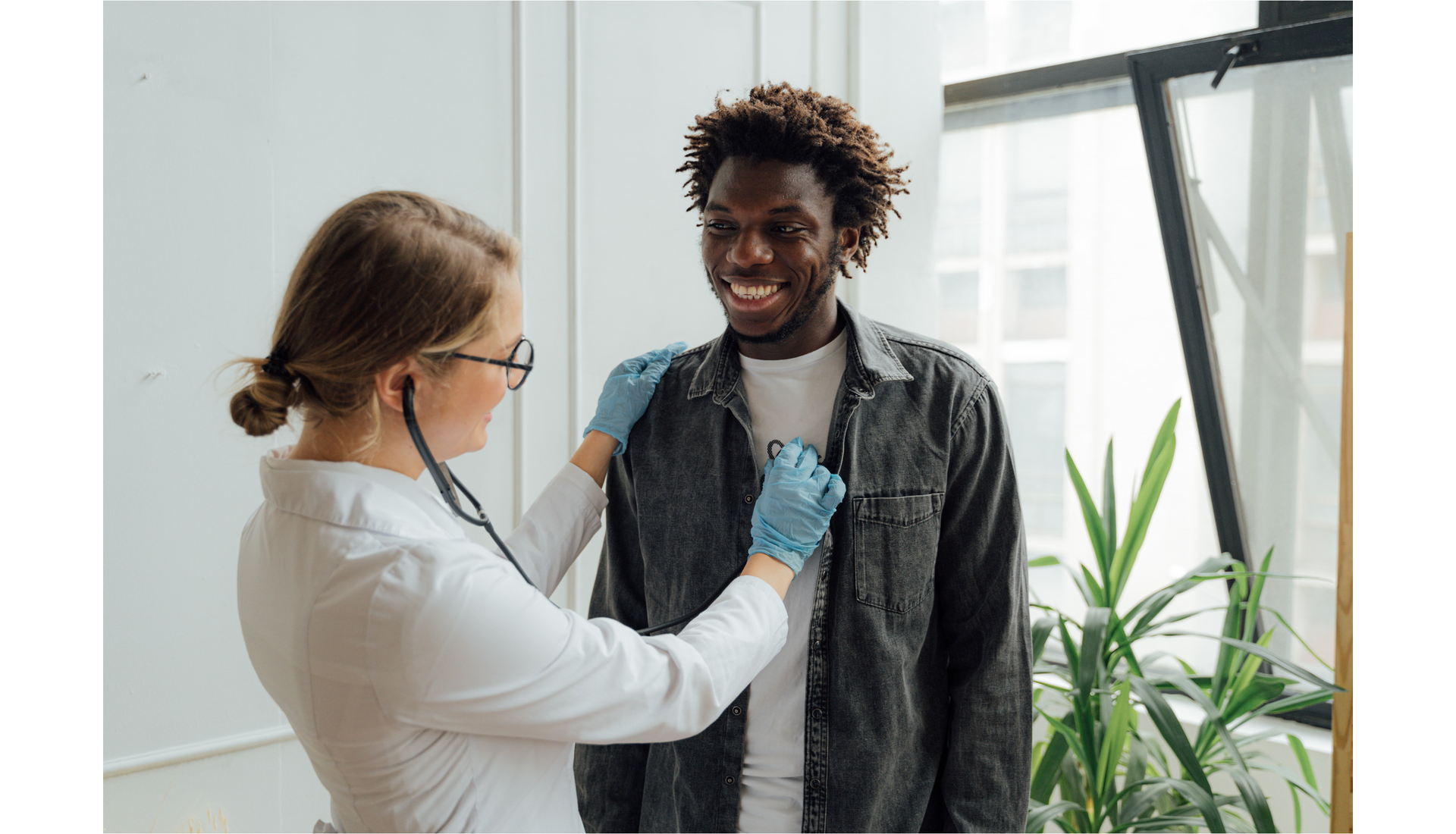 Zdjęcie przedstawia czarnoskórego, uśmiechniętego mężczyznę i kobietę w okularach w fartuchu lekarskim. Kobieta ma w uszach stetoskop. Kobieta stoi naprzeciwko mężczyzny i wykonuje badanie stetoskopem.