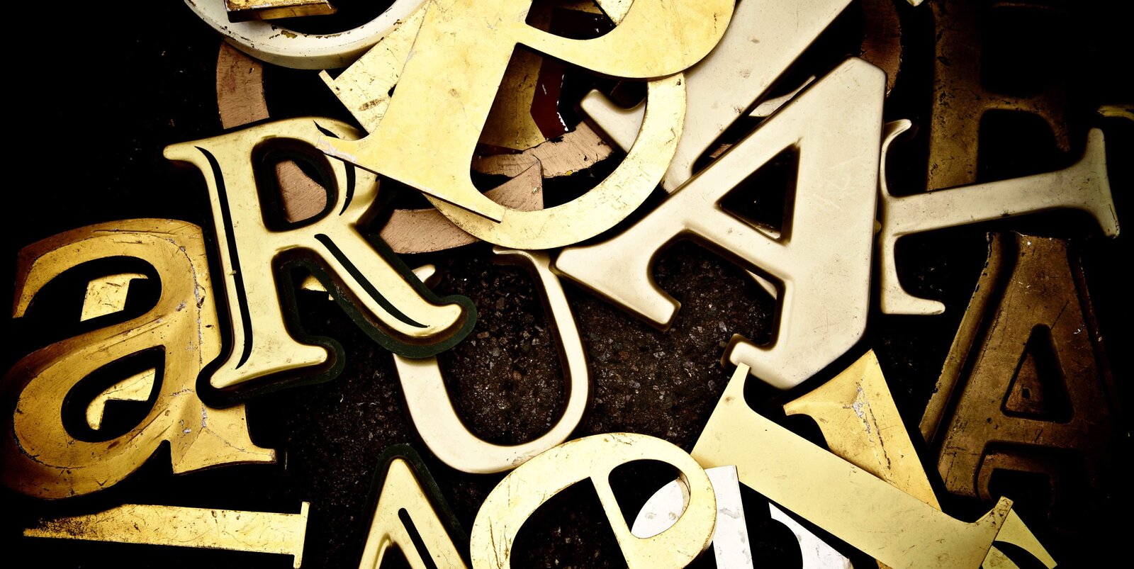 Fotografia przedstawia bezładnie rozrzucone na ciemnym tle małe i duże litery polskiego alfabetu, wykonane z metalu w złotym kolorze.