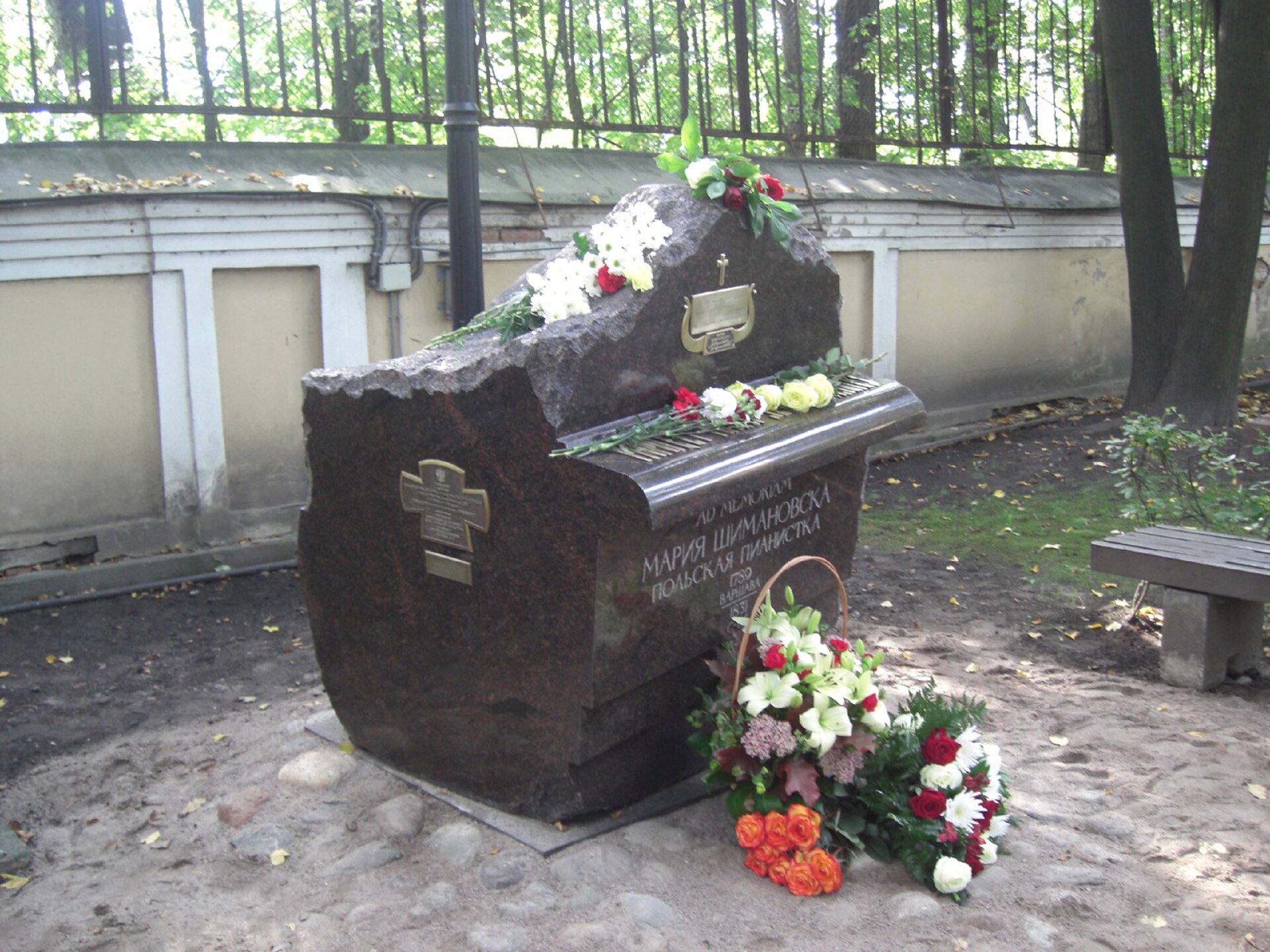 Ilustracja przedstawia ciemny pomnik Marii Szymanowskiej. Pomnik jest wysoki, zakończony niczym ułamana skała. Z przodu i z boku pomnika są krzyże. Przed pomnikiem i na pomniku leżą białe i czerwone kwiaty. 