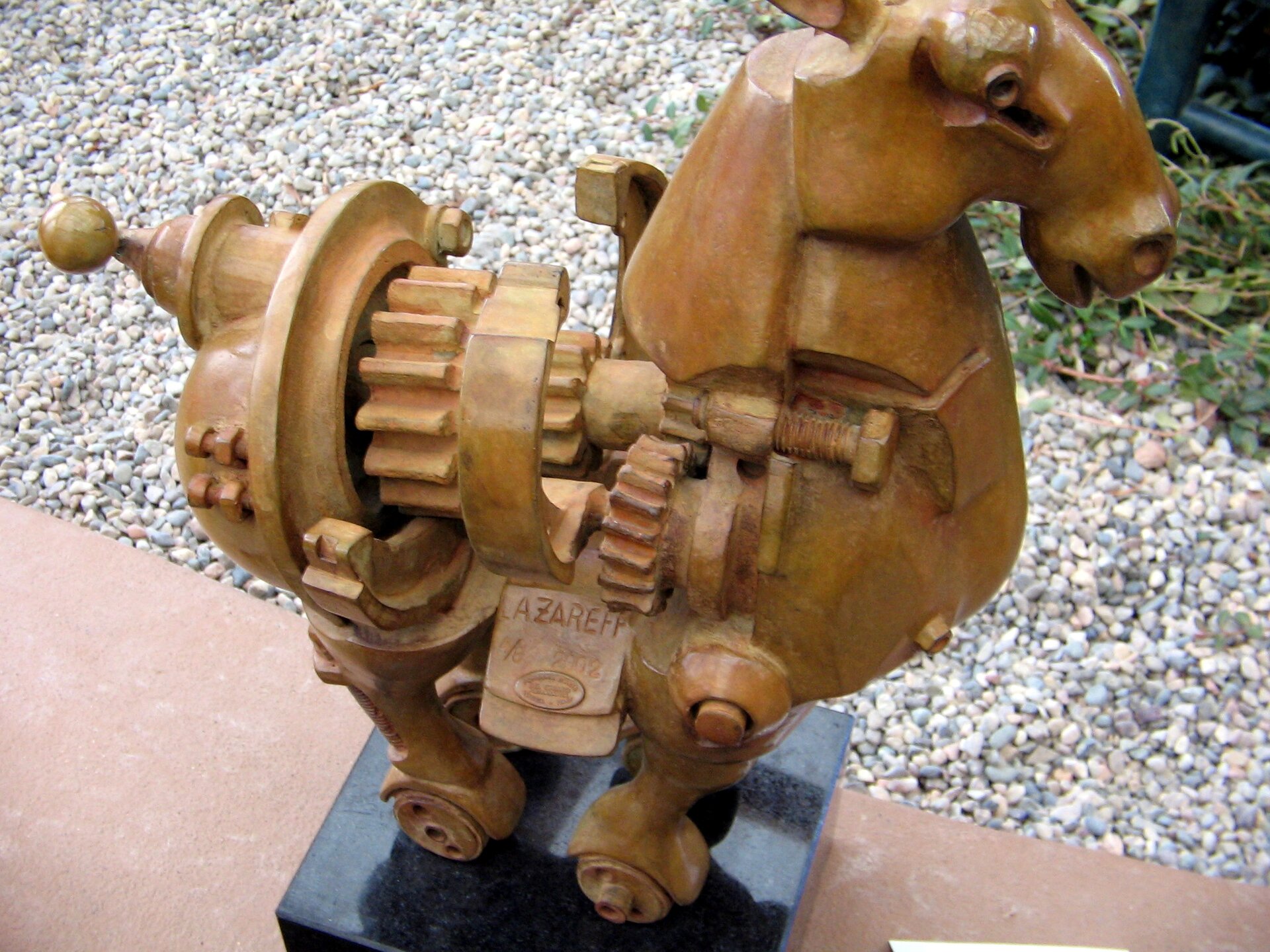 Fotografia przedstawia figurkę konia składającego się z wielu małych przekładni i innych mechanizmów. W tle ceglaste podłoże i żwirowy obszar z ubogą roślinnością.