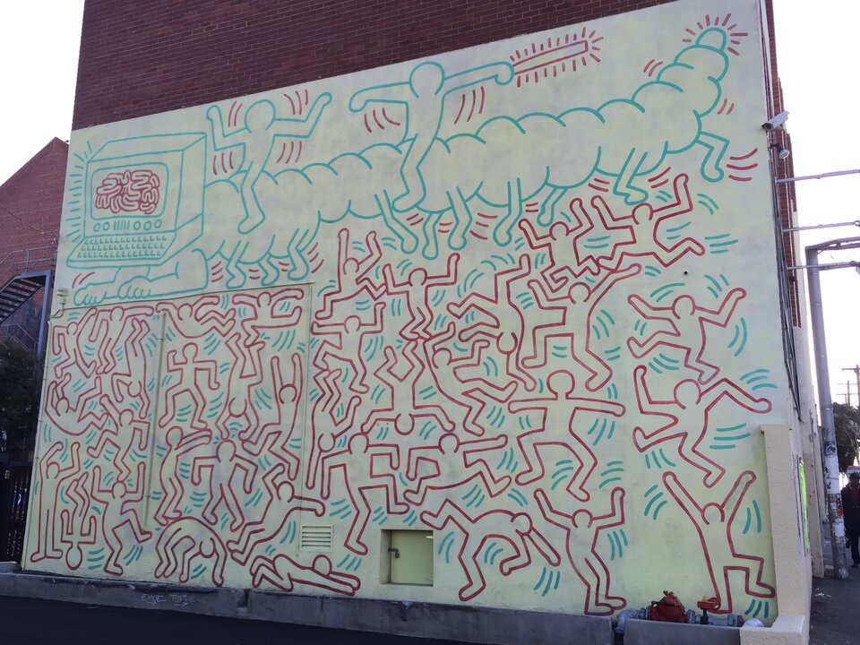 Fotografia przedstawia mural na ścianie budynku autorstwa Keith Haring. Ściana w białym kolorze pokryta jest płaskimi postaciami o czerwonych konturach, niewypełnionymi kolorem, są to schematyczne sylwetki. Postacie przybierają różne pozy, przeważnie jest to wyskok lub podskok. Nad nimi idzie gąsienica o niebieskich konturach, z komputerem zamiast głowy, na ekranie komputera przedstawiony jest czerwony mózg. Na grzbiecie gąsienicy jadą dwie postacie, jedna z nich trzyma podłużny przedmiot od którego odchodzą kreski przypominające promyki. Mural znajduje się na bocznej ścianie budynku z czerwonej cegły, w dolnej części ściany są małe beżowe, drzwiczki.