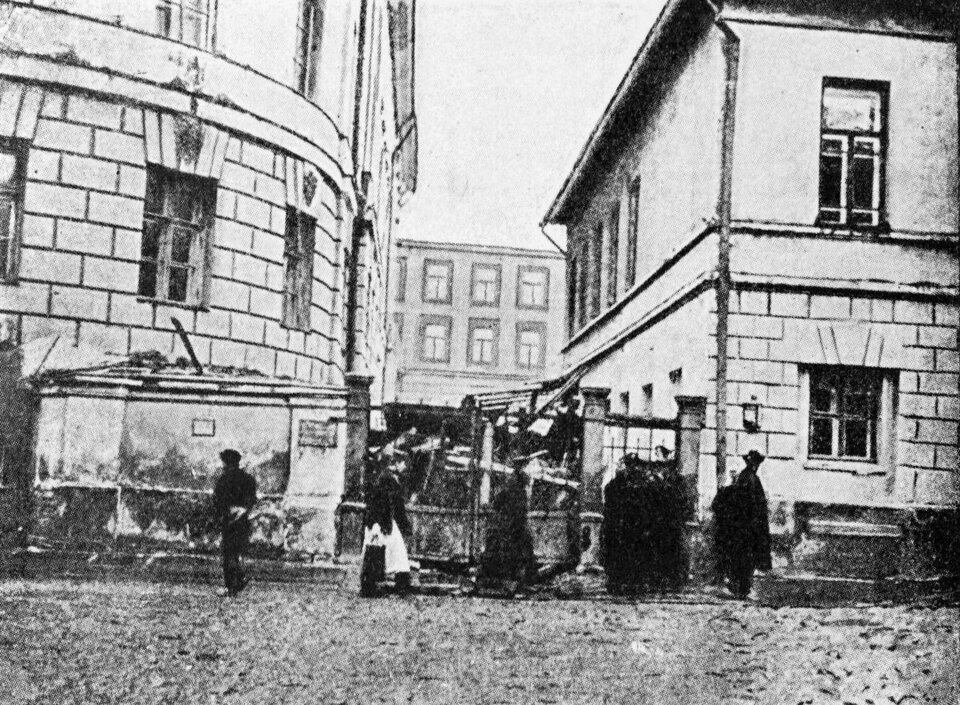 Fotografia przedstawia barykadę zbudowaną na ulicy pomiędzy dwoma kamienicami. Przed barykadą znajduje się brama. Widzimy ludzi idących wzdłuż ulicy.