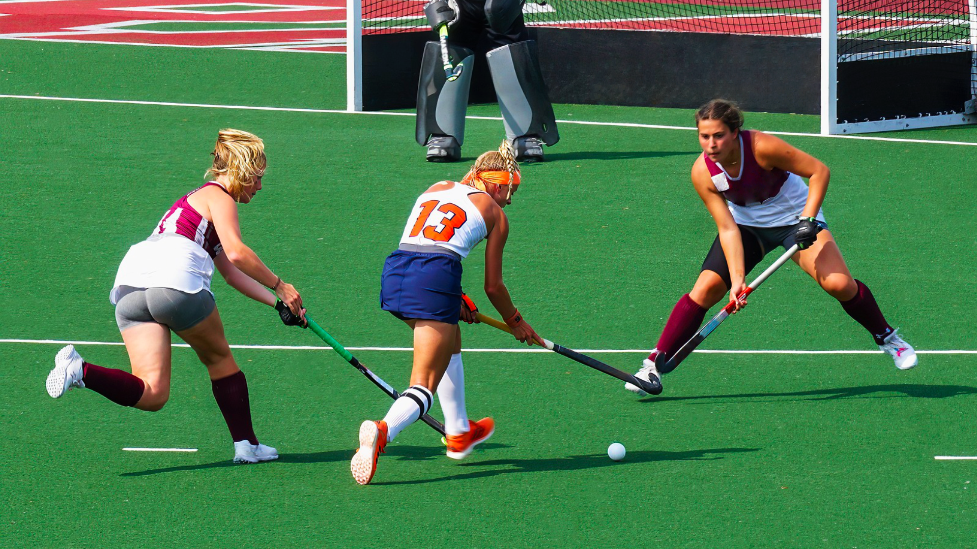 Zdjęcie przedstawia boisko z zieloną murawą, na którym dwie kobiety biegną do małej piłeczki. Kobieta z przeciwnej drużyny próbuje zatrzymać jedną z biegnących kobiet. W tle widać bramkę i osobę stojącą przed bramką z dużymi ochraniaczami na nogach. Wszystkie kobiety trzymają kije hokejowe.