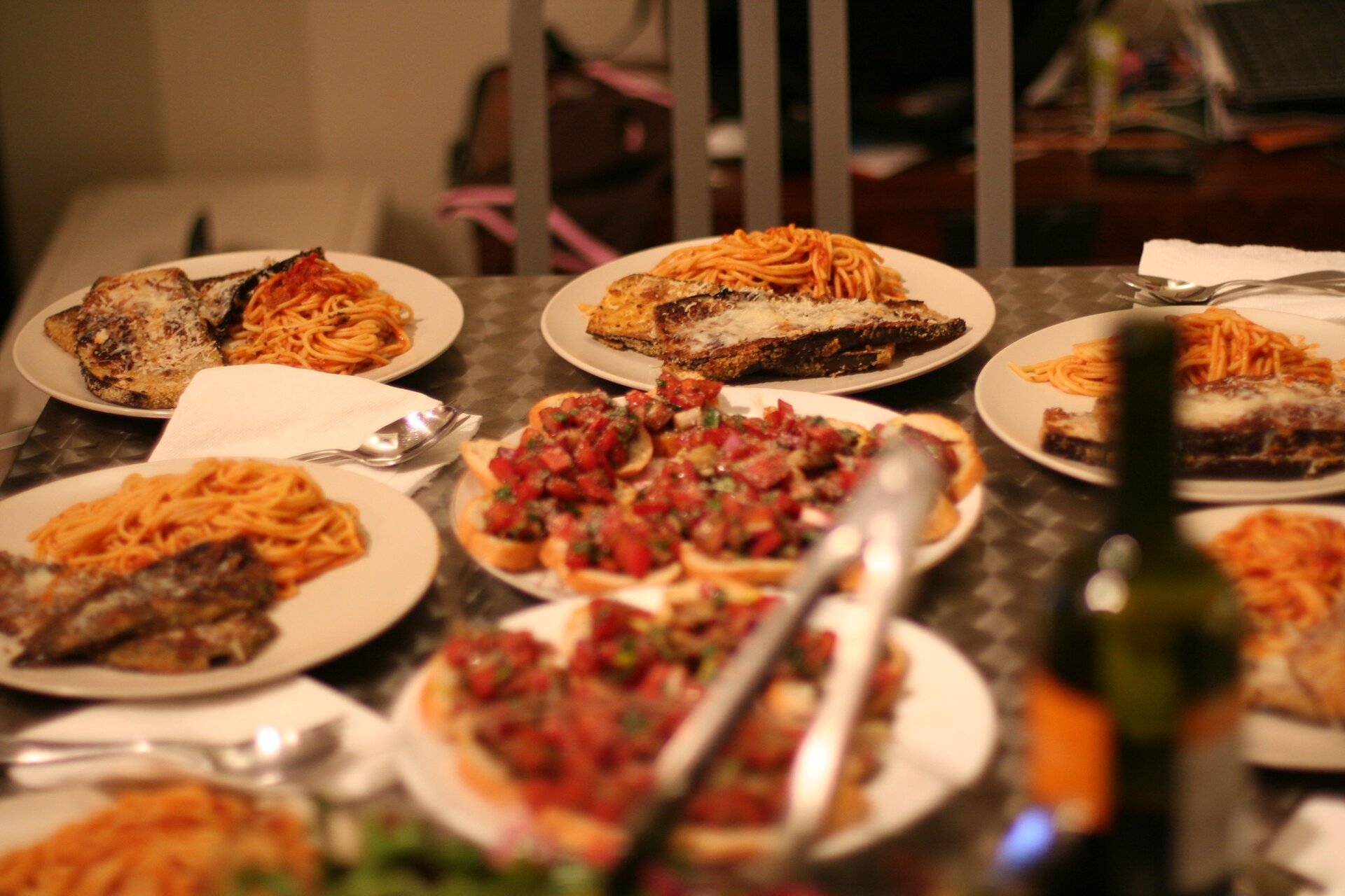 Kliknij aby powiększyć Na zdjęciu widać stół, na którym stoi sześć dużych talerzy z dużymi porcjami jedzenia oraz dwa duże talerze z przystawkami. Z przodu stoi butelka wina.