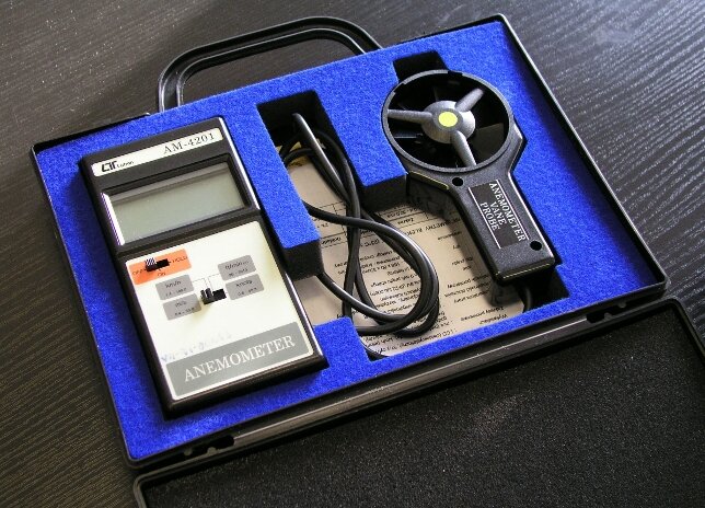 Anemometr wiatrakowy. Zdjęcie przedstawia urządzenie znajdujące się w otwartej walizce wyłożonej granatową pianką. Sprzęt składa się z trzech części. Pierwsza ma kształt prostokątnego urządzenia z małym ekranem, dwoma przyciskami i napisem: Anemometer. Obok znajduje się zwinięty czarny kabel. Trzecia część ma podłużny uchwyt zakończony okrągłą końcówką. W środku  jest pusta, ma tylko wirnik złożony z trzech ramion odchodzących od środka.