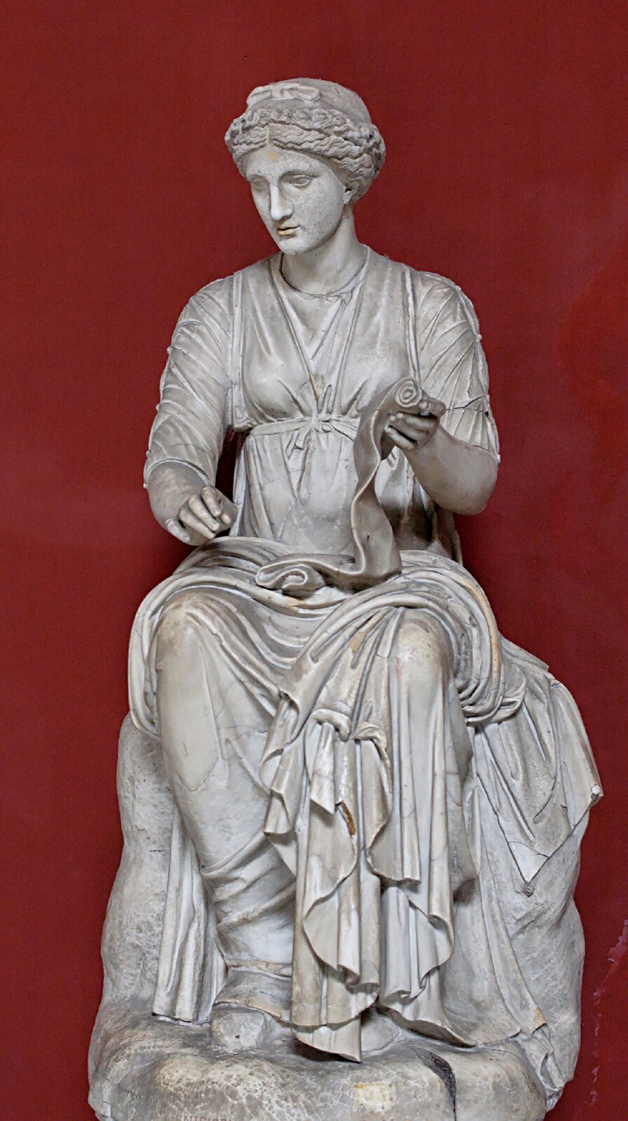 Ilustracja przedstawia rzeźbę Klio - muzy historii. Rzeźba wykonana jest z jasnego kamienia. Ukazuje siedzącą kobietę. Ma kręcone włosy. Ubrana jest w długą szatę. W lewej dłoni trzyma zwój papirusu. Prawa dłoń uniesiona jest nad lewym udem. Rzeźba ustawiona jest na tle bordowej ściany.