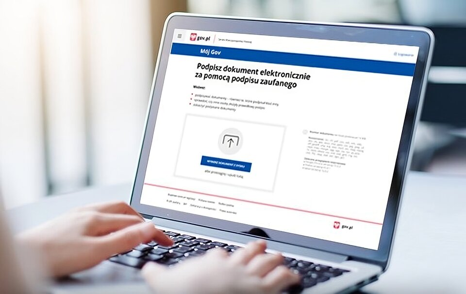 Ilustracja przedstawia komputer przenośny, na którym wyświetlona jest strona internetowa serwisu ePUAP.  W lewym górnym rogu widać godło Polski a obok niego napis: gov.pl. Poniżej znajduję sie napis: Podpisz dokument elektronicznie za pomocą podpisu zaufanego, i krótka instrukcja jak to zrobić.  Widoczne są dłonie osoby, która wpisuje coś na klawiaturze. 