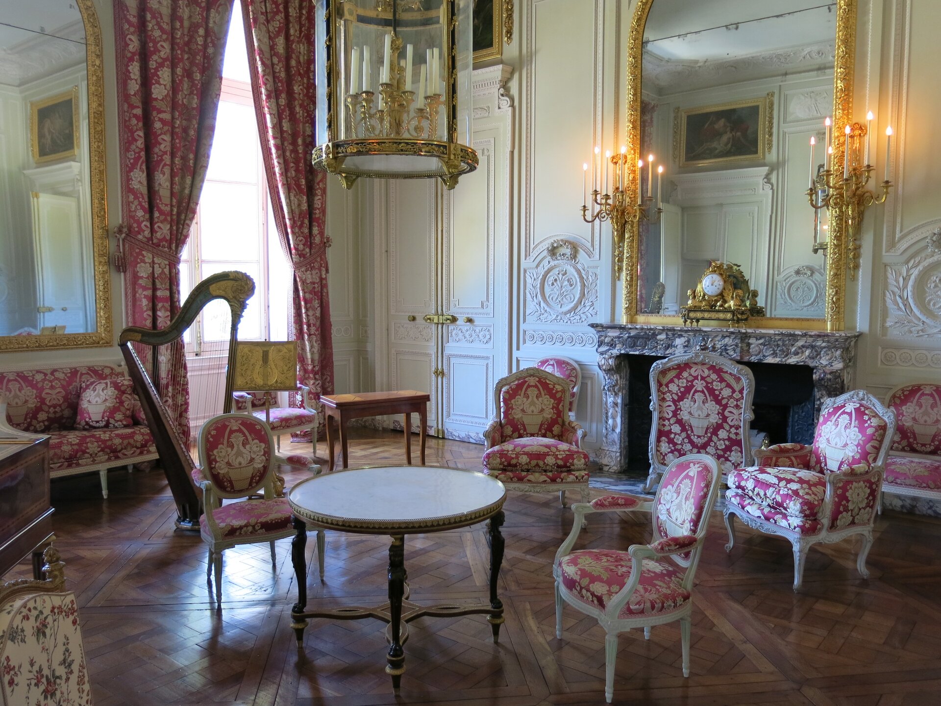 Ilustracja przedstawia zdjęcie wnętrza salonu urządzonego w stylu rokoko. Na środku salonu stoi niewielki okrągły stół. Obok stołu stoją dwa fotele. Nad stołem wisi żyrandol ze świecami. Po prawej stronie stoją dwa większe fotele. Za nimi znajduje się kominek, nad którym wisi duże lustro w złotej, ozdobnej ramie. Po bokach lustra wiszą dwa złote kinkiety. Na kominku stoi złoty zegar. W głębi zdjęcia znajdują się białe, dwustronne drzwi, obok drzwi znajduje się wysokie okno, na którym wisi kotara. Kotara jest w kolorze bordowym z białym haftem. Taką samą tkaniną obite są fotele i sofa znajdująca się obok okna. Nad sofą wisi również duże lustro w złotej ramie. Przed sofą stoi harfa. Obok harfy stoi prostokątny, drewniany, mały stolik, obok niego stoi fotel. Podłoga w salonie zrobiona jest z ciemnego drewna. Ściany w salonie są białe, zdobione białą sztukaterią. 