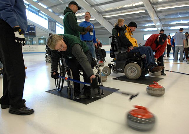 Zdjęcie przedstawia grupkę ludzi na lodowisku, dwoje z przodu siedzą na wózkach inwalidzkich, jedna z osób jest pochylona do przodu i  trzyma w ręku kij do curlingu którym popycha granitowy kamień. 