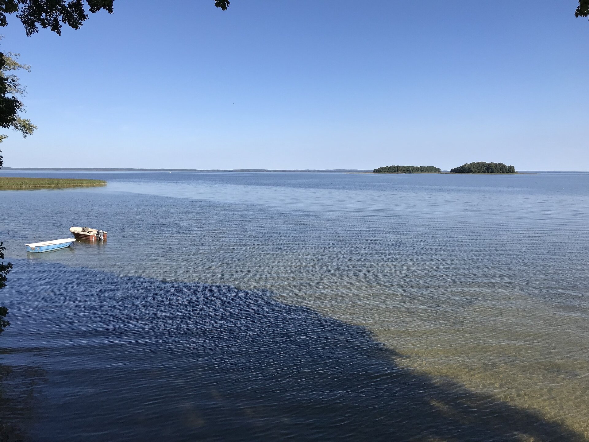 Fotografia bardzo rozległego jeziora. Dookoła w oddali brzegi pokryte drzewami. Na pierwszym planie niewielkie łódki.