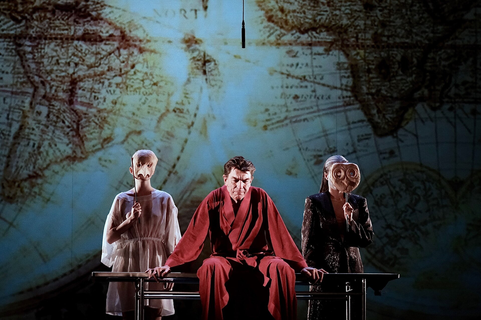 Zdjęcie przedstawia kadr ze spektaklu. Na pierwszym planie fotografii ukazany jest mężczyzna, ubrany w czerwony szlafrok. Jego wzrok skierowany jest w dół. Mężczyzna siedzi na ławce. Po jego prawej i lewej stronie znajdują się dwie kobiety w maskach. Kobieta po lewej stronie, ubrana jest w białą, przeźroczystą sukienkę. Kobieta po prawej stronie, ubrana jest w czarną sukienkę. Drugi plan tworzy tło, ukazujące mapę. 