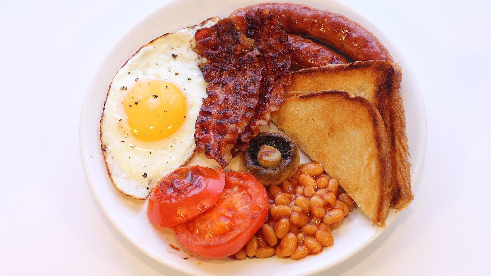 Na zdjęciu widoczny jest biały, okrągły talerz z jedzeniem. Obok sadzonego jajka podano pieczone plasterki pomidora, wstążki pieczonego bekonu, kiełbaski, fasolę w sosie pomidorowym, tosty przekrojone w trójkąty, a na środku upieczoną pieczarkę.