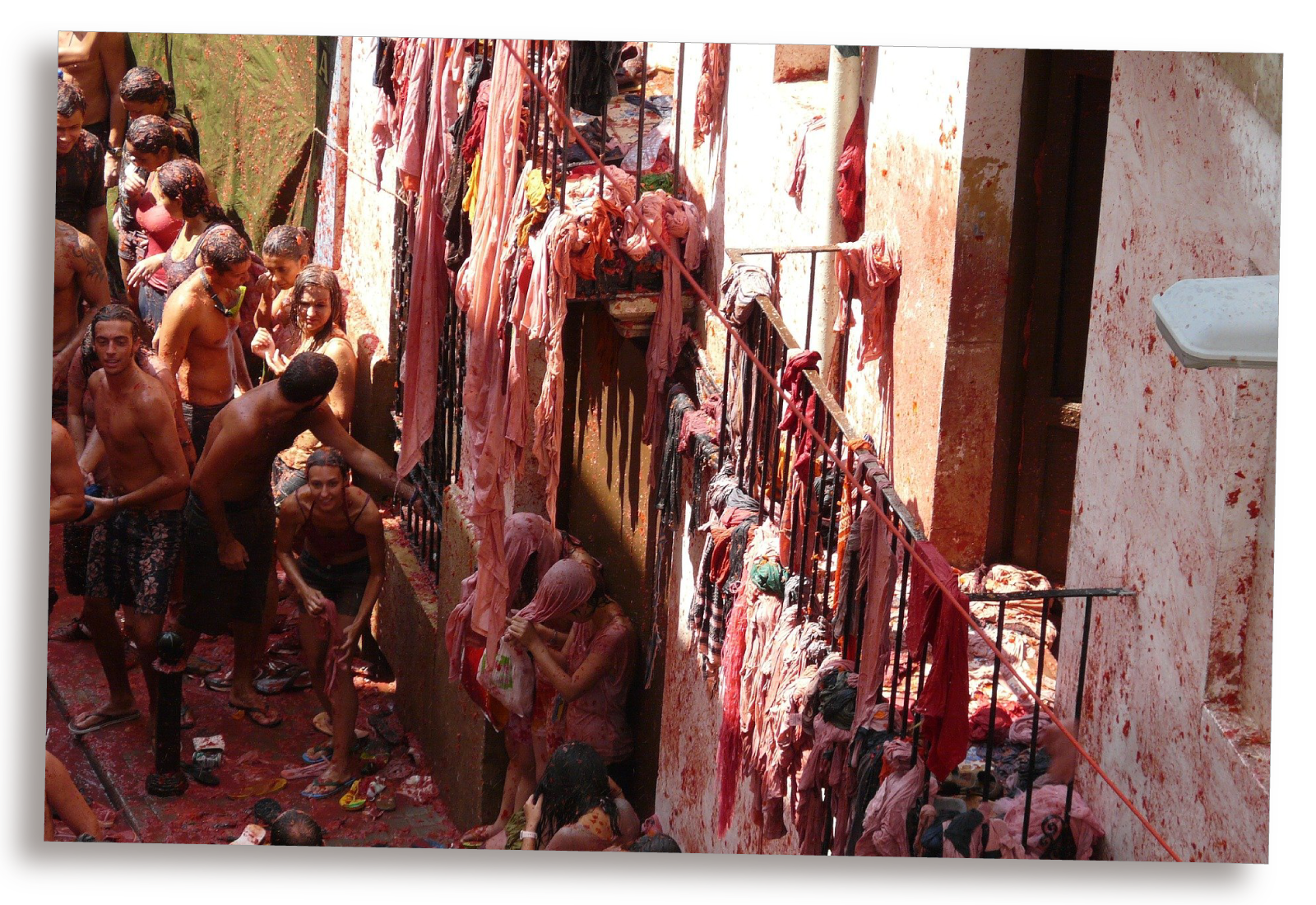 Zdjęcie przedstawia uliczkę ubrudzoną pomidorami pełną ludzi bez koszulek. Z balkonów zwisają materiały.