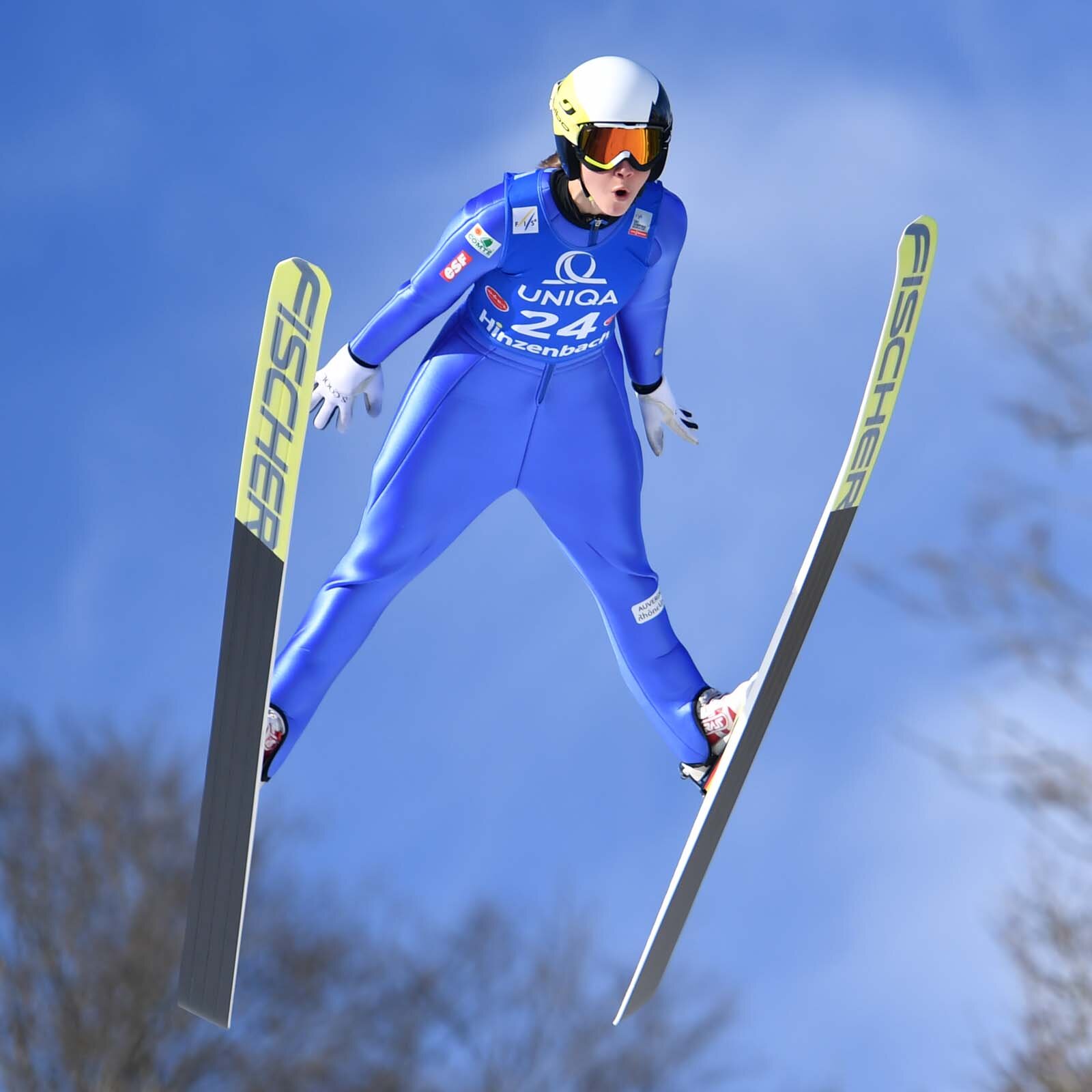 Zdjęcie przedstawia sportowca w trakcie wykonywania skoku na nartach. Ujęcie zrobione jest od dołu. Mężczyzna ubrany jest w kombinezon oraz w kask i gogle.