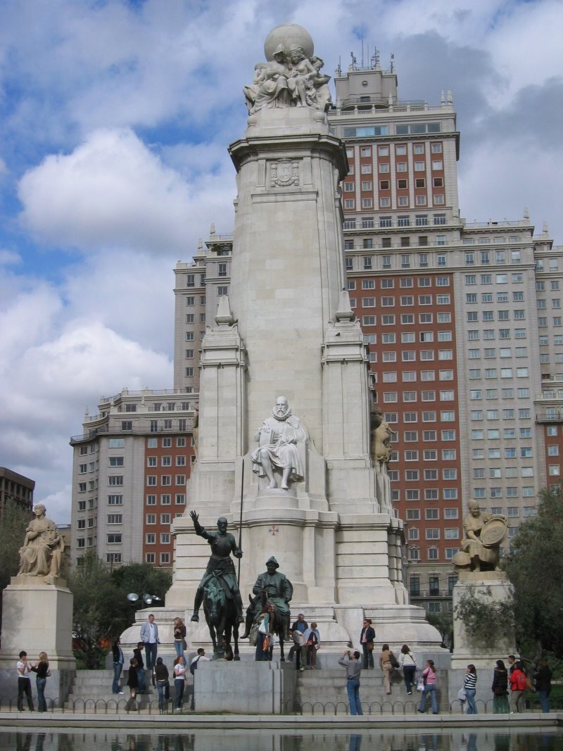 Kliknij, aby powiększyć Pomnik Miguela de Cervantesa . Głównym elementem pomnika jest obelisk. To kamienna, biała kolumna. Na jej szczycie znajduje się glob ziemski otoczony przez pięć kobiet. U stóp obelisku, na cokole, znajduje się siedząca postać Miguela de Cervantesa wykonana z kamienia. Don Kichot na koniu i Sancho Pansa na ośle stoją przed posągiem pisarza, figury są wykonane z brązu. Po bokach obelisku znajdują się rzeźby z piaskowca przedstawiające dwie wizje ukochanej rycerza: prostą dziewczynę Aldonzę Lorenzo oraz piękną, wyobrażoną Dulcyneę z Toboso. W tle znajduje się wysoki, ponad dwudziestopiętrowy budynek. Wokół pomnika stoją ludzie, przyglądają się monumentowi i robią zdjęcia.
