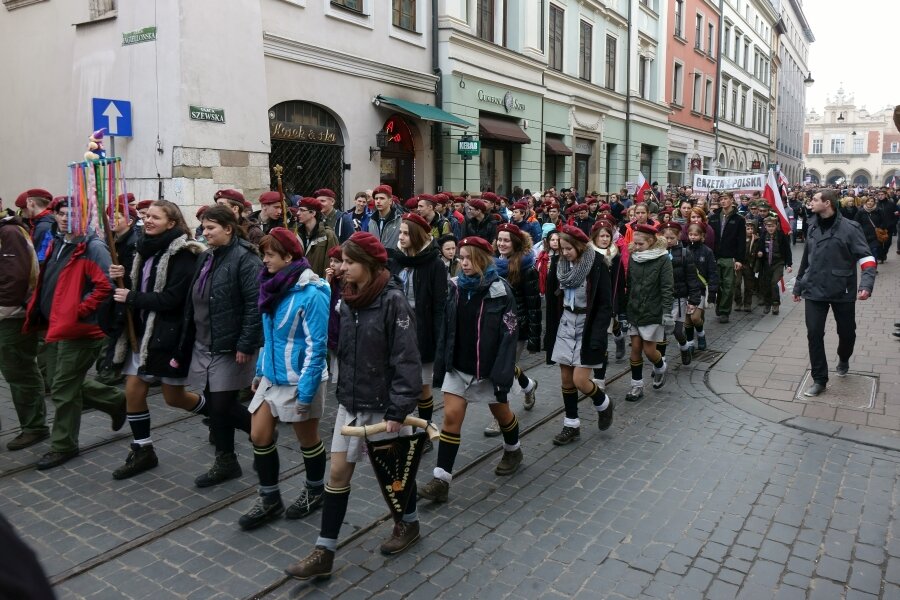 Zdjęcie przedstawia pochód idący ulicami miasta. W pochodzie biorą udział harcerze i harcerki ubrani w mundury i fioletowe berety. Niosą flagi Polski i transparenty.