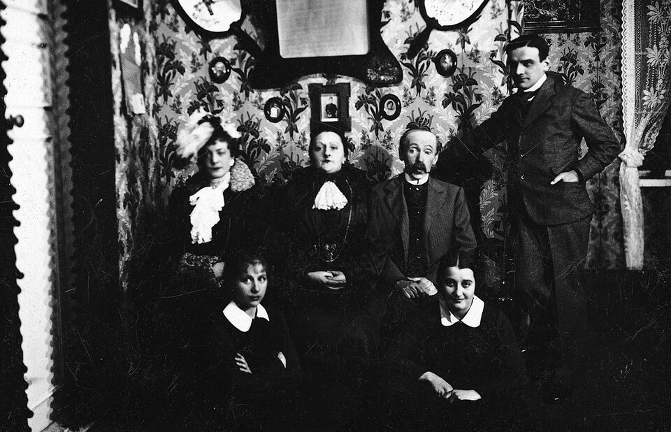 Zdjęcie przedstawia grupę sześciu osób pozujących w pomieszczeniu o wzorzystych ścianach, na których zawieszone są liczne obrazy i fotografie w ozdobnych ramach. W centralnej części siedzi kobieta w ciemnym stroju z jasnym żabotem, a po jej lewej stronie mężczyzna z wąsami w garniturze. Po bokach i u ich stóp rozmieszczone są pozostałe osoby: stojący mężczyzna po prawej, siedząca kobieta w kapeluszu z piórami po lewej oraz dwie młode kobiety siedzące na podłodze na pierwszym planie.