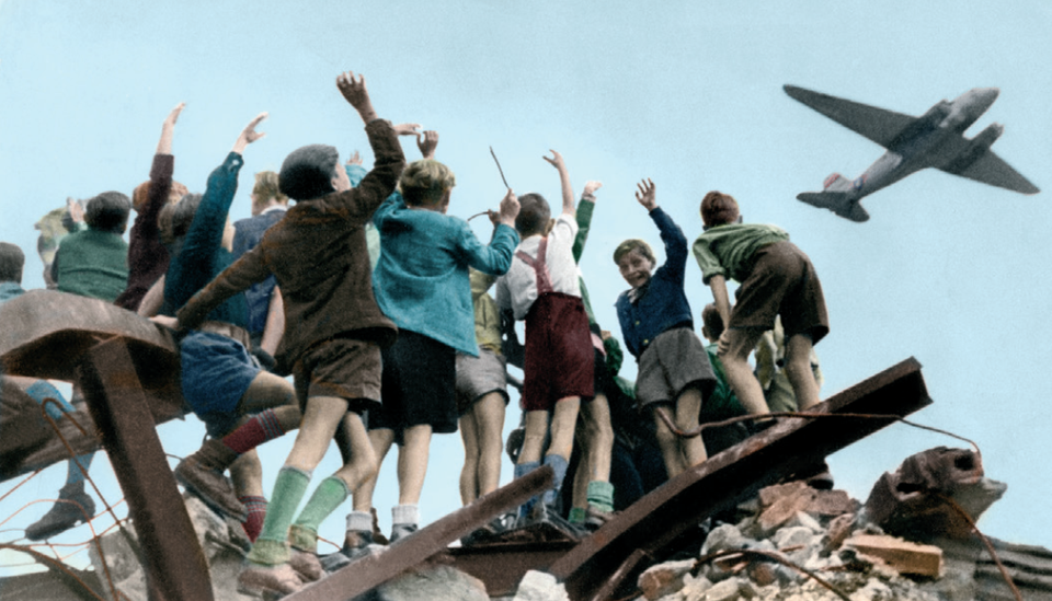 Zdjęcie przedstawia lecący samolot. Do samolotu macha grupa chłopców, która stoi na rumowisku.