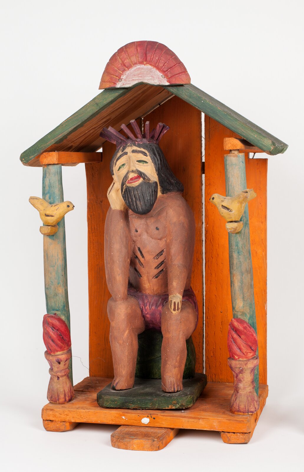 Zdjęcie przedstawia wykonaną z drewna kapliczkę. Jest to rzeźba siedzącego na kamieniu Jezusa, podtrzymującego głowę na prawej ręce. Ma on czarne włosy oraz czarny zarost. Oczy ma prawie zamknięte, a usta - lekko uchylone. Na jego głowie jest korona cierniowa. Nad nim znajduje się drewniany daszek, pomalowany na zielono, podtrzymywany na dwóch nóżkach. Na nóżkach wyrzeźbiono wizerunki ptaków. 