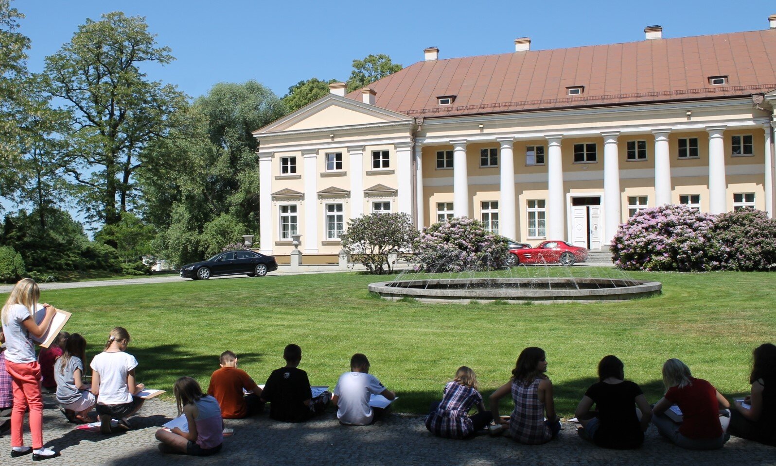 Zdjęcie przedstawia biało‑żółty budynek, którego przednia część jest podparta na kolumnach. Przed budynkiem znajdują się niskie krzewy, a także zielona trawa. Wokół budynku, z lewej strony zdjęcia, rosną wysokie, zielone drzewa. Na trawie siedzą uczniowie ubrani w kolorowe ubrania, którzy pochylają się nad kartkami, po których piszą. Niebo jest błękitne, bezchmurne. Zdjęcie zostało wykonane w świetle dziennym.