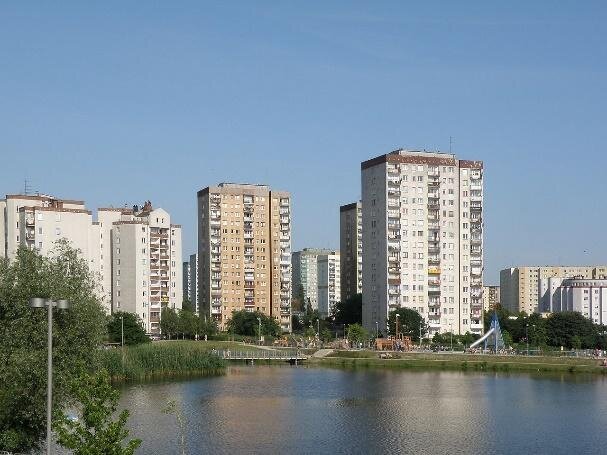 Zdjęcie przedstawia osiedle składające się z wysokich bloków. Wokół nich są pasy zieleni, na których rosną drzewa i trawa. Przed nimi znajduje się zbiornik wodny. 