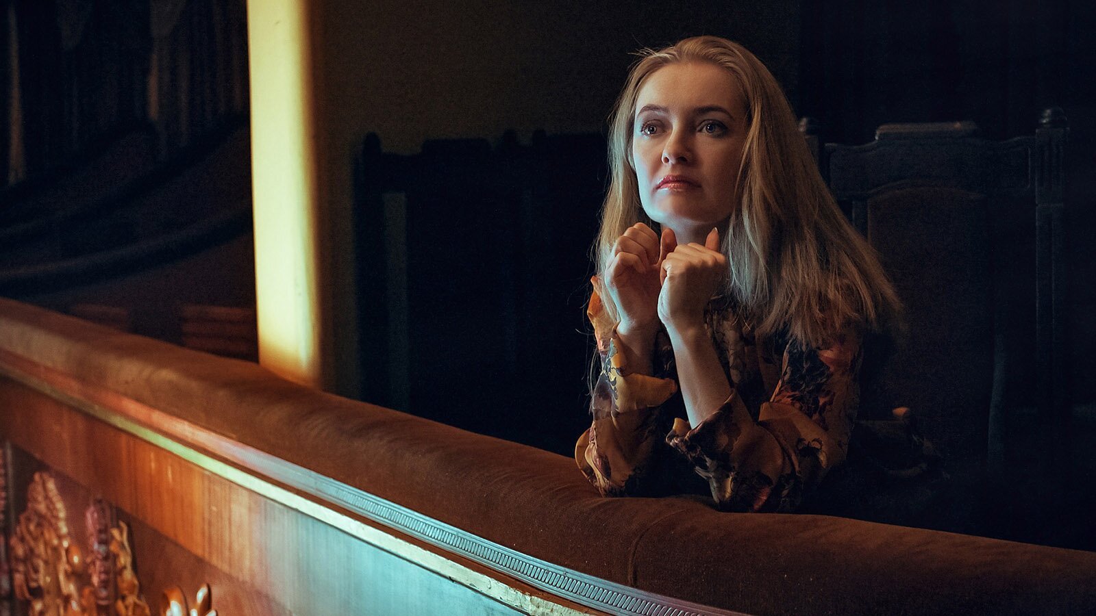 Zdjęcie przedstawia młodą kobietę, w ujęciu od pasa w górę, stojącą na balkonie w teatrze. Kobieta ma długie, jasne włosy i jest ubrana we wzorzystą bluzkę. Spogląda w górę. Ręce ma zgięte w łokciach, oparte na drewnianej balustradzie, a dłonie zaciśnięte w pięści.