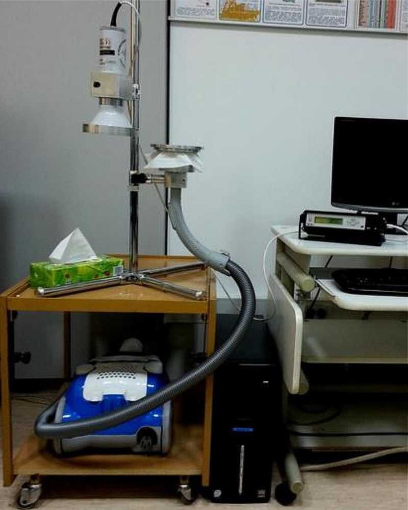 Rys. a. Rysunek przedstawia zdjęcie układu służącego do pomiaru stężenia gazu promieniotwórczego – radonu. Składa się on z odpowiednio przystosowanego odkurzacza z rurą wyprowadzoną pionowo do góry zakończoną lejkiem z odpowiednim systemem filtrującym. Oprócz tego układ ten zawiera zawieszony na pionowym wysięgniku skierowany w dół czujnik pomiarowy w postaci szarego walca zakończonego u dołu także lejkiem. Czujnik ten podłączony jest do miernika i komputera dokonujących pomiaru.