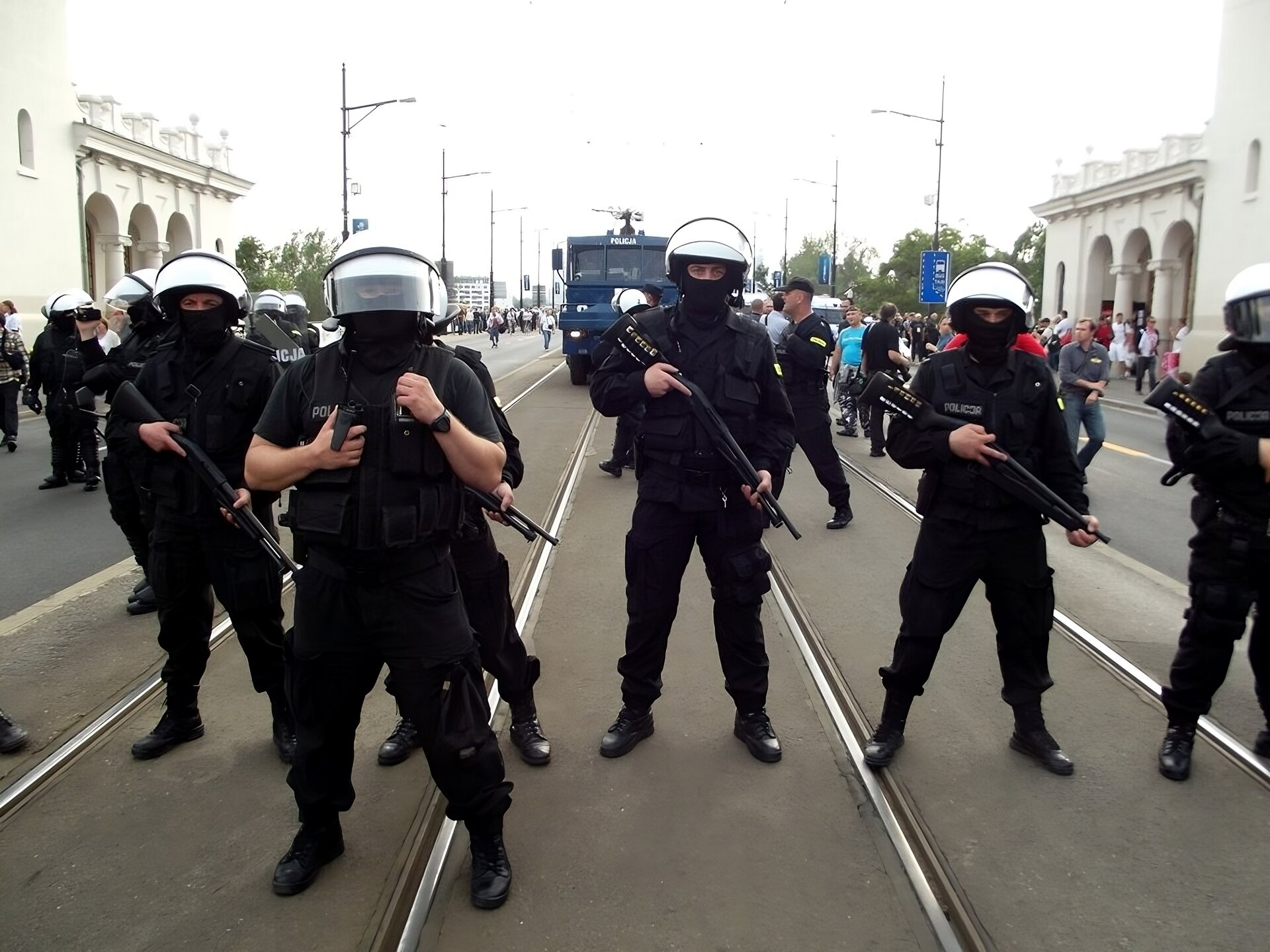 Zdjęcie, które przedstawia blokadę policyjną.  Policjanci ustawieni w szeregu na ulicy. Za nimi widoczni ludzie i auto policyjne. Policjanci ubrani w mundur ochronny, widoczną kamizelka ochraniającą z napisem policja. Na głowie założony kask ochronny z osłoną zabezpieczająca twarz. Dodatkowym zabezpieczeniem twarzy policjantów są ubrane kominiarki. Stojący w rozkroku policjanci trzymają w rękach broń długą.Po prawej i po lewej stronie budynki. 
