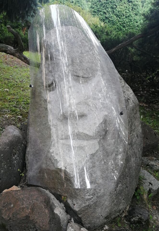 Ilustracja przedstawia fotografię rzeźby plenerowej. W kadrze znajduje się rzeźba wykonana w kamieniu i plastiku. Przedstawia twarz, która jest przesłonięta przezroczystą, półokrągłą osłoną.