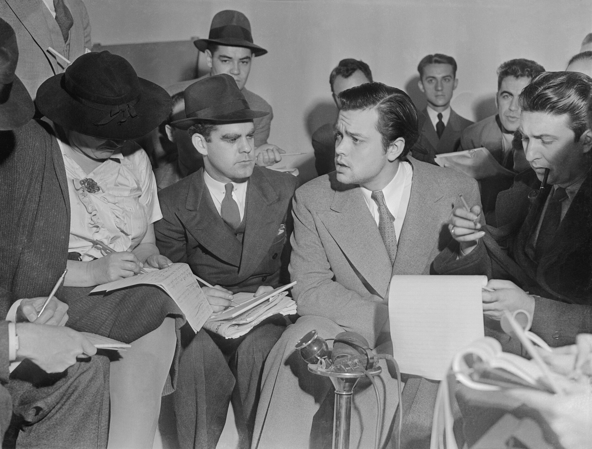 Zdjęcie przedstawia grupę młodych mężczyzn w garniturach oraz jedną kobietę, którzy siedzą ściśnięci obok siebie. Trzymają notesy i notują. Jeden z mężczyzn przemawia. Stoi przed nim mikrofon.