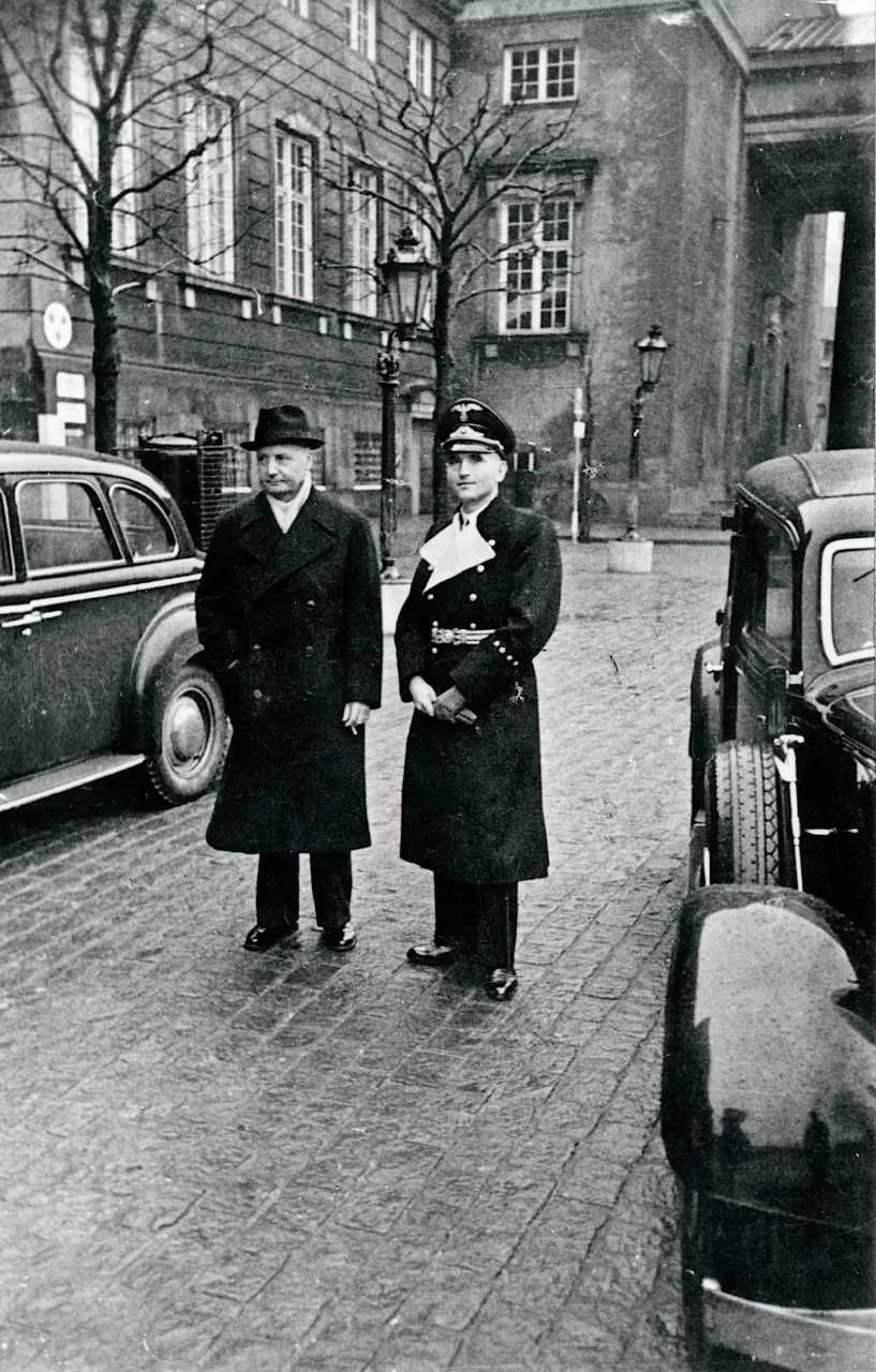 Zdjęcie przedstawia mężczyzn. Stoją oni na wybrukowanej ulicy. Mężczyzna z lewej ma kapelusz, czarny płaszcz i białą koszulę pod spodem. Drugi ma czapkę oficerską, płaszcz spięty pasem i z odgiętym kołnierzem. Mężczyźni znajdują się pomiędzy zaparkowanymi samochodami. W tle znajdują się nagie drzewa, a za nimi wysoki budynek.