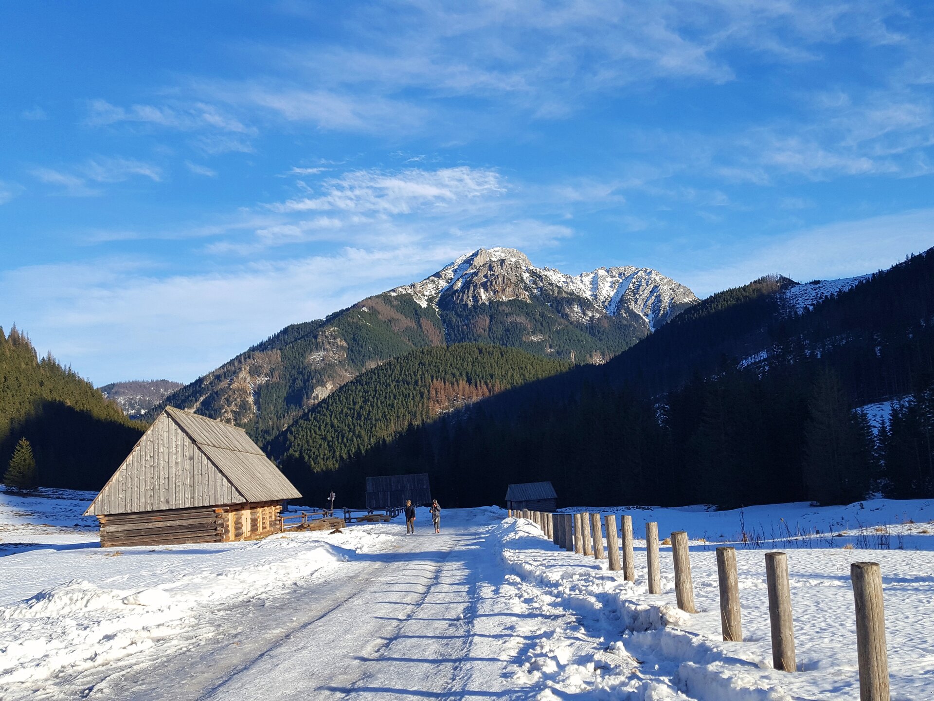 Na fotografii widoczna jest Dolina Chochołowska w zimowym krajobrazie. Na pierwszym planie ukazana jest ośnieżona ścieżka, po której podążają dwie osoby. Po bokach znajdują się dwie, niewielkie, drewniane, jasnobrązowe chatki góralskie. Na drugim planie widoczne jest szare, porośnięte ciemnym lasem, wysokie pasmo górskie.