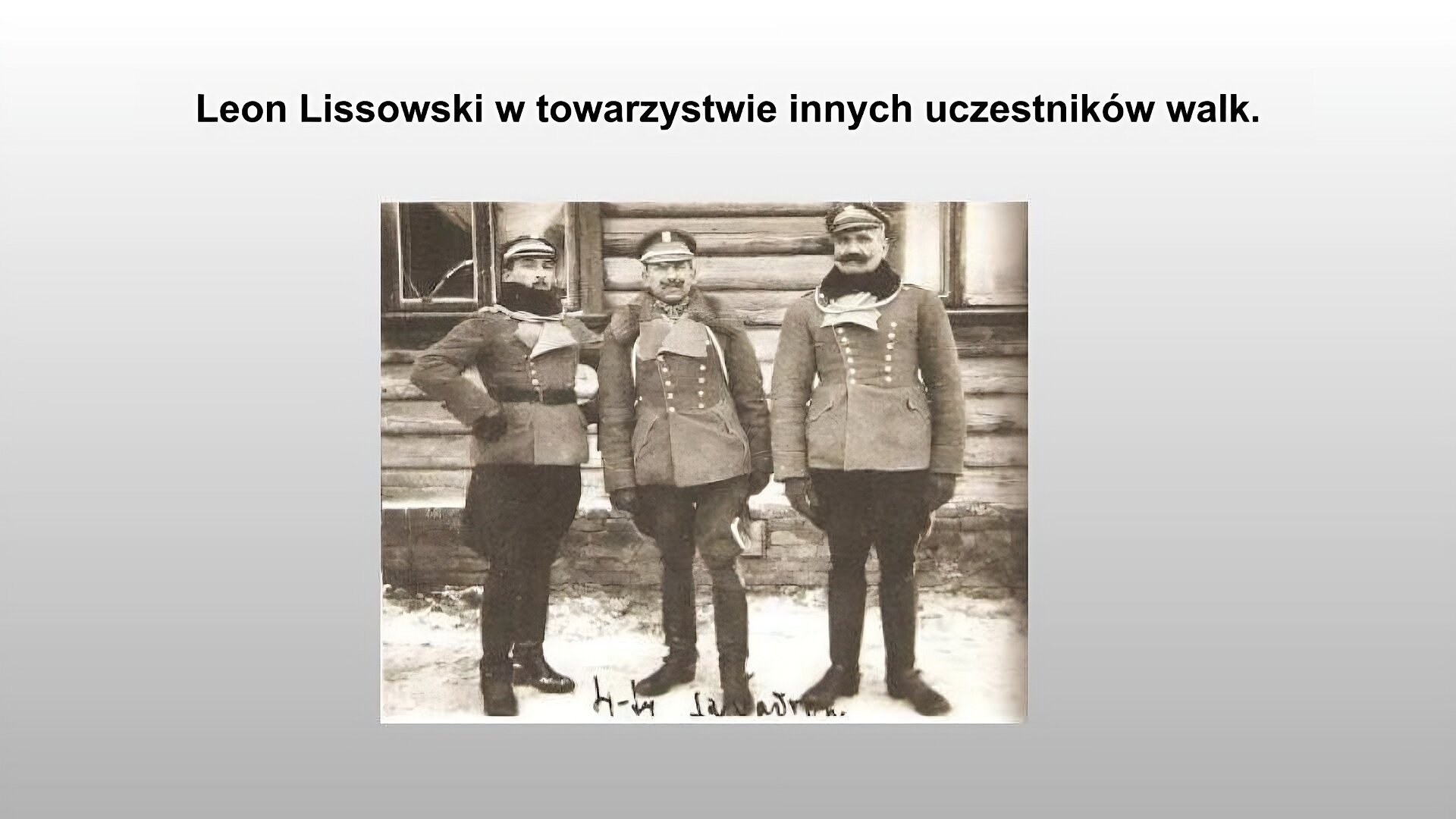 Jasnoszary slajd. Na górze slajdu tytuł: „Leon Lissowski w towarzystwie innych uczestników walk”. Poniżej zdjęcie przedstawiające trzech mężczyzn. Ubrani są podobnie - każdy z nich ma ciemne spodnie, przylegające do łydek i swobodnie zwisające na udach. Każdy z nich ma kurtkę zapinaną na guziki, której kołnierze są białe. Mężczyzna po prawej i lewej stronie mają na szyjach czarne szaliki. Wszyscy mężczyźni mają na głowach czapki wojskowe. Mężczyźni stoją na tle drewnianego domu. Na ziemi leży śnieg. Zdjęcie zostało wykonane w sepii.