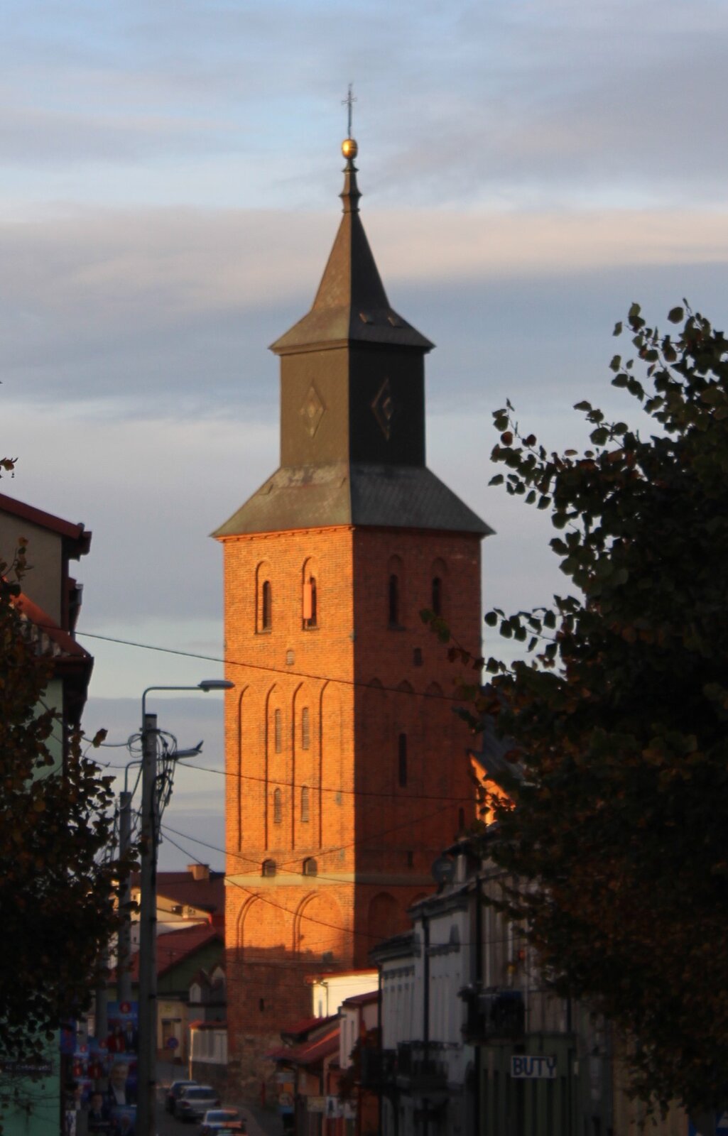 Zdjęcie przedstawia wysoką wieżę wykonaną z czerwonej cegły. Na jej szczycie znajduje się ciemny, wysoki dach, na szczycie którego widoczny jest krzyż. Wieża znajduje się przy ulicy, przy której znajdują się kolorowe kamienice. Wzdłuż ulicy stoją zaparkowane samochody.