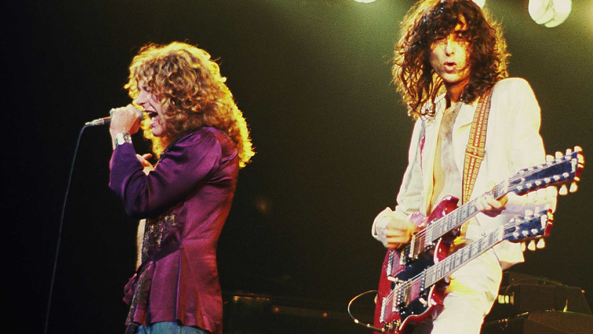 Ilustracja przedstawia zespół Led Zeppelin - brytyjska grupa założona w Londynie, będąca jednym z pionierów hard rocka. Mężczyźni znajdują się na scenie podczas koncertu. Jeden z nich śpiewa, natomiast drugi gra na gitarze. Mężczyźni mają długie, kręcone włosy. Wokalista ma fioletową marynarkę, drugi białą.