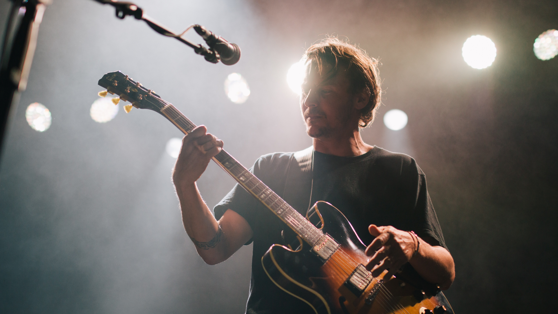 Zdjęcie przedstawia artystę na scenie z gitarą elektryczną. Obok widoczny jest mikrofon na statywie.