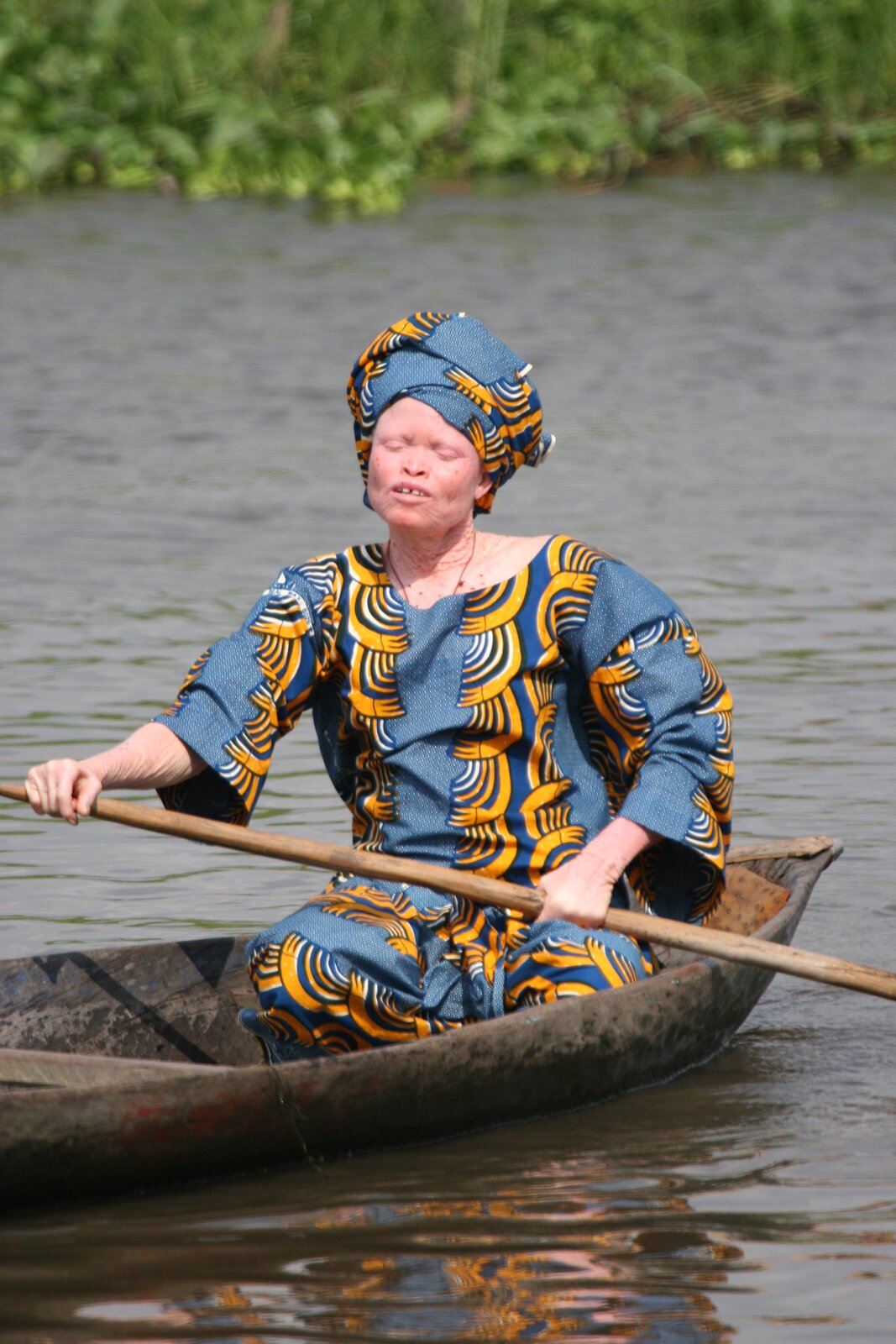 Zdjęcie przedstawia kobietę w barwnej sukience i barwnym turbanie na głowie, która płynie małą łodzią po rzece. Kobieta białą skórę z wyraźnie widocznymi zmarszczkami i pofałdowaniami. Nie ma brwi ani rzęs. Jej oczy są zamknięte. 