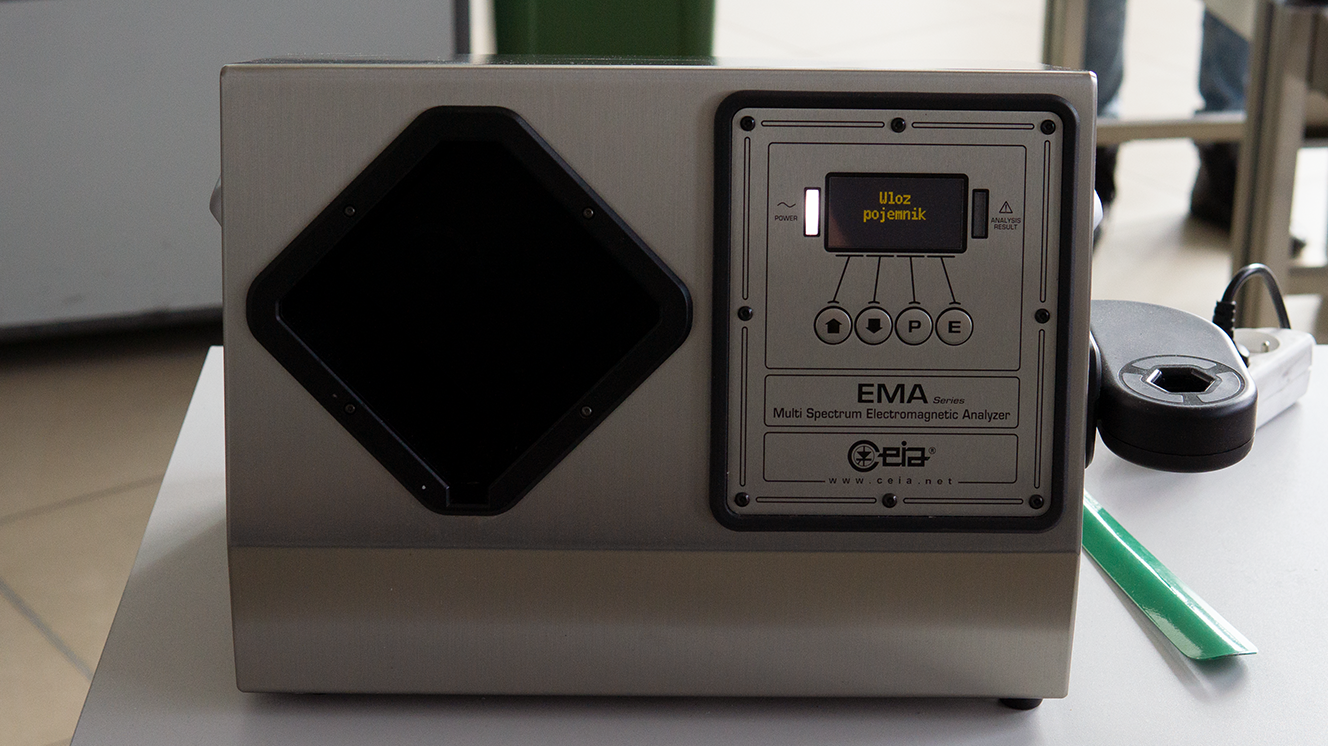 Zdjęcie przedstawia eL E De Se. w nawiasie likłid eksplosif detekszyn system. czyli system do wykrywania materiałów niebezpiecznych w płynach, aerozolach lub żelach. Jest to małe, szare urządzenie. Leży ono na biurku. Po lewej stronie przyrządu jest czarny romb. Po prawej jest ramka, a w nie od góry jest ekranik z napisem. Włóż pojemnik. Z ekraniku wychodzą na dół cztery strzałki do czterech przycisków. Na pierwszym przycisku jest strzałka w górę, na drugim jest strzałka w dół, na trzecim litera Pe, a na czwartym litera E. po bokach ekraniku są dwa światełko. Lewe światełko świeci, a obok niego jest napis. Pałer. Prawa światełko nie świeci, a obok niego jest nieczytelny angielski napis. Poniżej jest tekst. ema siries. Multi spektrum elektromagnetik analizer. Pod spodem jest logo c e i a oraz adres strony. Wu wu wu kropka ce e i a kropka pe el. Z prawej strony urządzenia wystaje czarny element z dziurą. Dziura ma kształt rombu. Za przyrządem na biurku leżą zielona łyżka do butów oraz przedłużacz z jedną włożoną wtyczką.