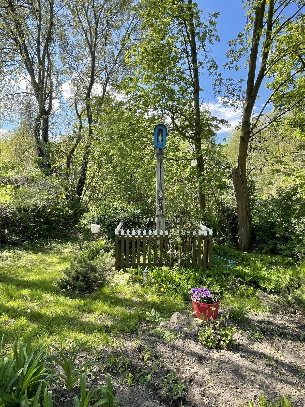 Fotografia przedstawia kapliczkę. Wokół niej zarośla i drewniany płotek, wewnątrz którego rosną kępiaste kwiaty. Za kapliczką drzewa. Pośrodku terenu kapliczki odgrodzonego płotkiem stoi jasny, drewniany słupek o wysokości nieco ponad dwóch metrów, na którym umieszczono niebieską , pionową skrzynkę z zaokrąglonym daszkiem, wewnątrz której znajduje się niewielka figurka Matki Bożej.