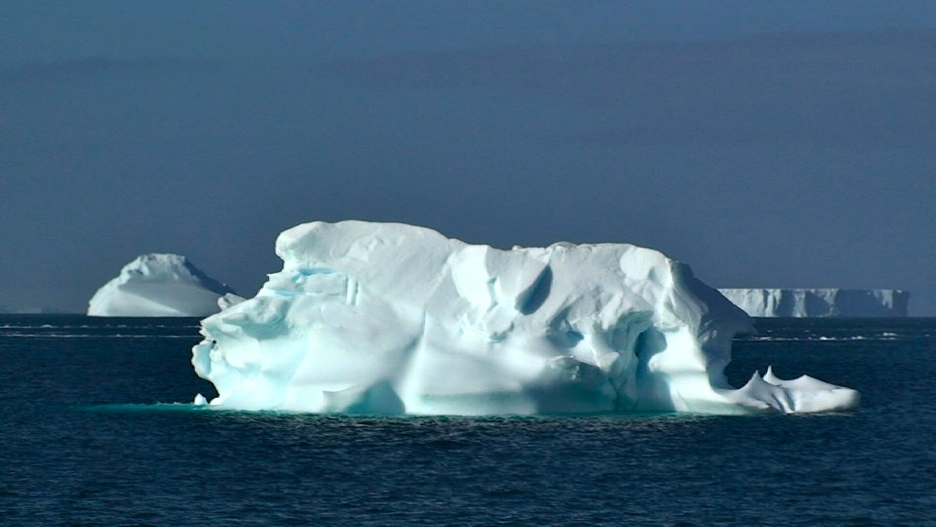 Na zdjęciu na pierwszym planie góra lodowa w wodzie. W tle zbita masa lodu w wodzie oraz ciemnoniebieskie niebo.