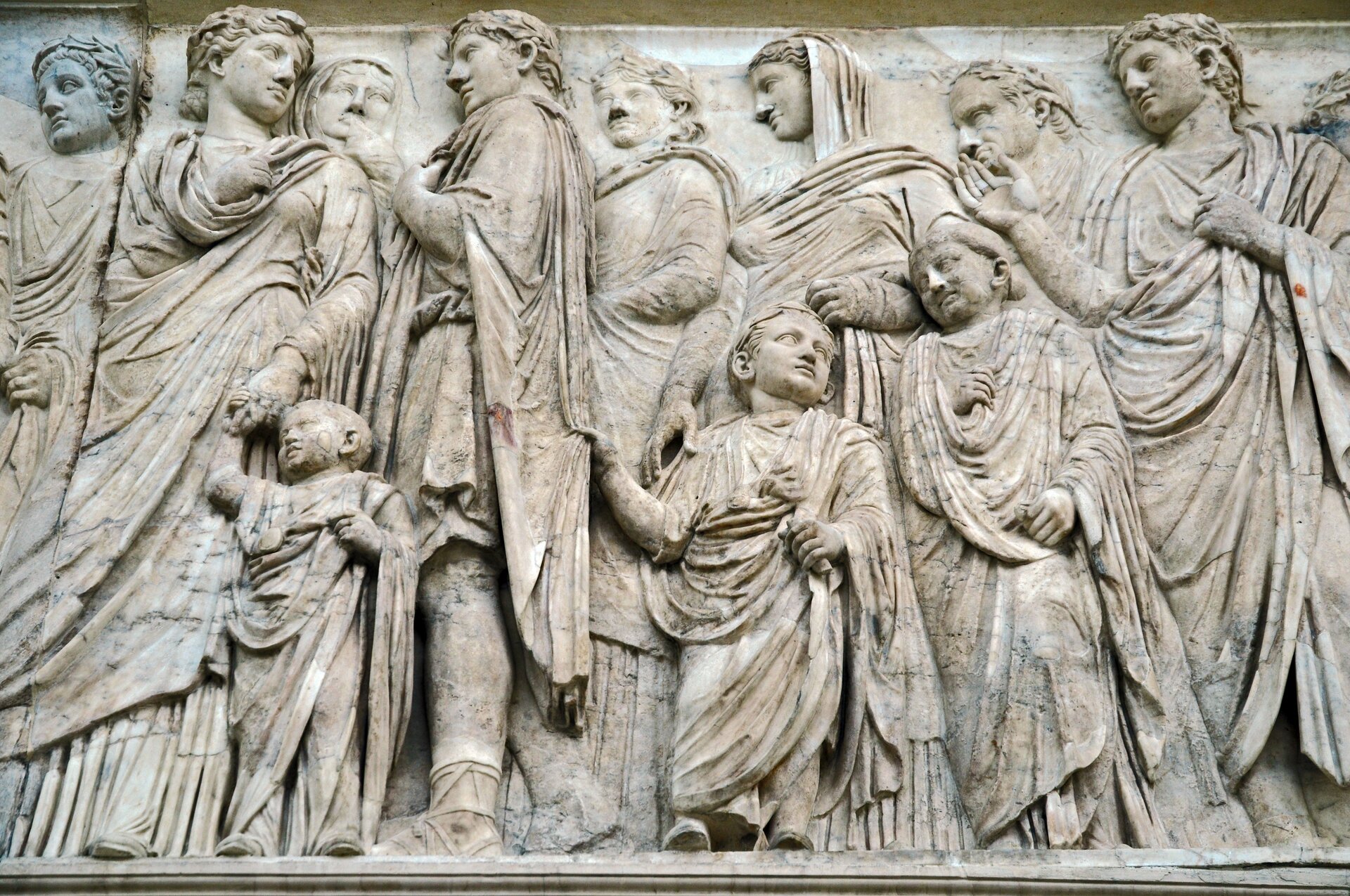 Fotografia przedstawia płaskorzeźbę, która ukazuje grupę osób. Większość z nich ma głowy odwrócone w bok. Grupa składa się z dorosłych i dzieci. Wszyscy ubrani są w długie togi.